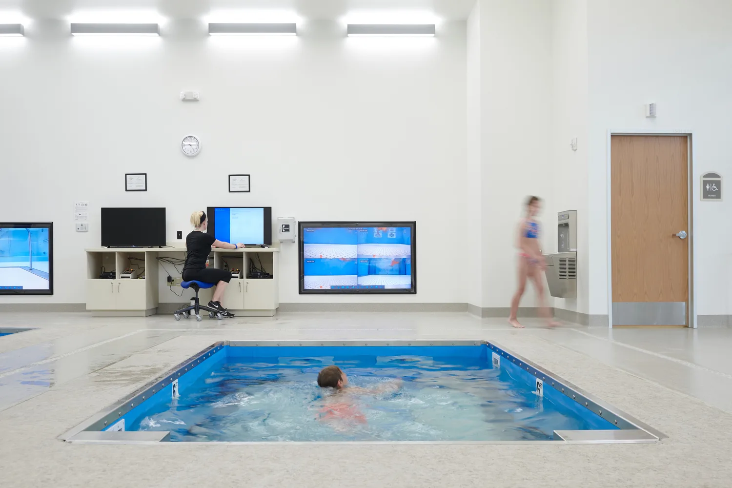 An indoor sports training and rehabilitation facility with two small interior pools for exercise and physical therapy. A woman in exercise clothing sits at a large computer surrounded by TV screens showing an underwater view of the pool to monitor patient movement. A child is swimming in the pool. The high-ceiling space features white hexagonal acoustic panels and industrial-style lighting. This photo demonstrates BSA's expertise in architecture, engineering, and designing Sports Medicine facilities.