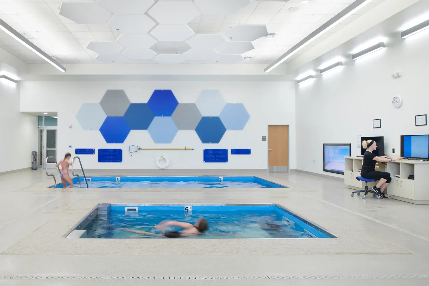 An indoor sports training and rehabilitation facility with two small interior pools for exercise and physical therapy. A woman in exercise clothing sits at a large computer surrounded by TV screens showing an underwater view of the pools to monitor patient movement. One child is swimming in the closer pool, while another child is descending down a ladder into the pool in the background. The high-ceiling space features white hexagonal acoustic panels and industrial-style lighting. This photo demonstrates BSA's expertise in architecture, engineering, and designing Sports Medicine facilities.