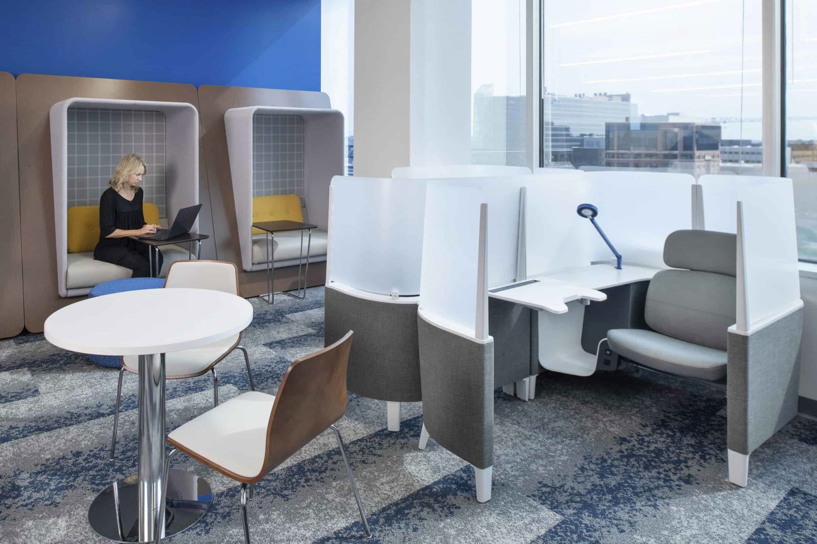 Various workspace options in a modern office featuring multiple privacy solutions. On the left, a person with blonde hair in black clothing works on a laptop within a pod-like enclosure with gray checkered interior backing and yellow cushioning, designed for focus work with sound absorption. In the foreground, a small round white table with wooden chairs offers a casual meeting spot. To the right, individual workstations with white and gray privacy screens provide semi-private desk areas with ergonomic chairs. Large windows reveal city views, and blue patterned carpeting continues the color scheme seen throughout the space. The design showcases different levels of privacy and work preferences in a post-pandemic flexible office environment, from fully enclosed pods for concentration to semi-private workstations and collaborative areas.
