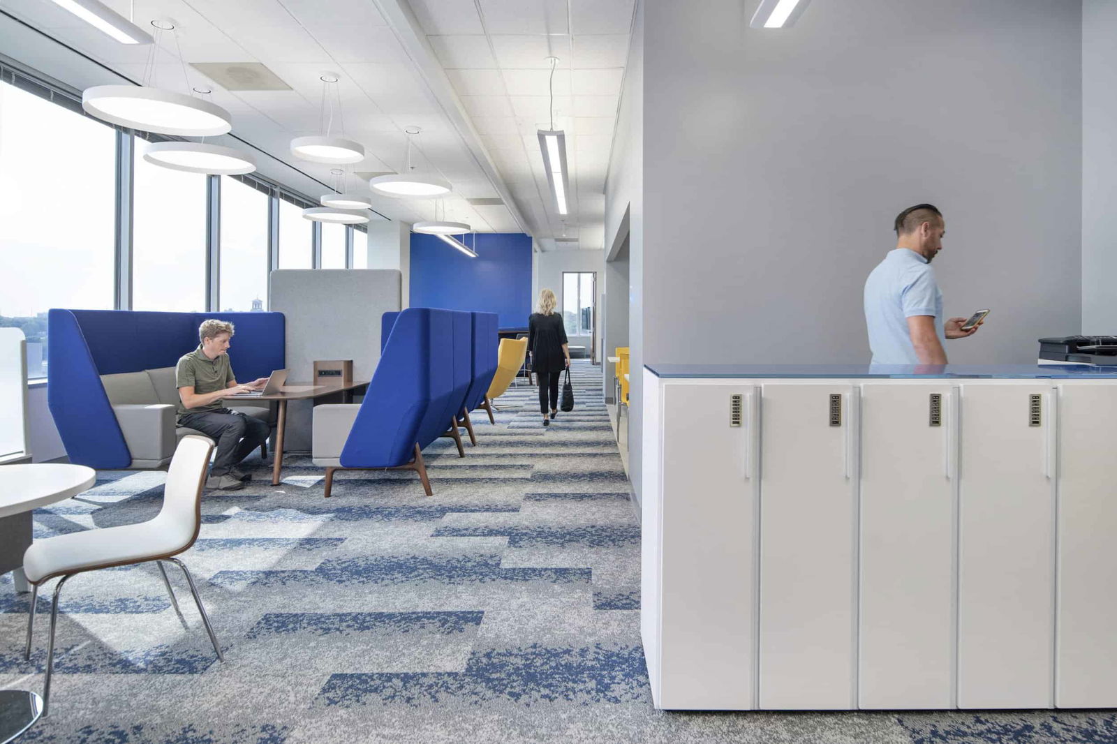 Open office layout featuring multiple work zones with a consistent blue and white color scheme. On the left, a person in a green shirt works on a laptop in a semi-private blue booth with high backs that provide acoustic privacy while remaining open to the environment. In the center walkway, a person in black clothing walks toward the back of the office. To the right, a person in a light blue shirt checks their phone while standing at a counter area, adjacent to a row of white storage lockers with numbered locks. The space features modern circular pendant lighting, large windows with city views, and blue patterned carpeting. The design demonstrates post-pandemic workplace solutions that combine personal storage options with various semi-private work areas while maintaining an open, collaborative atmosphere.