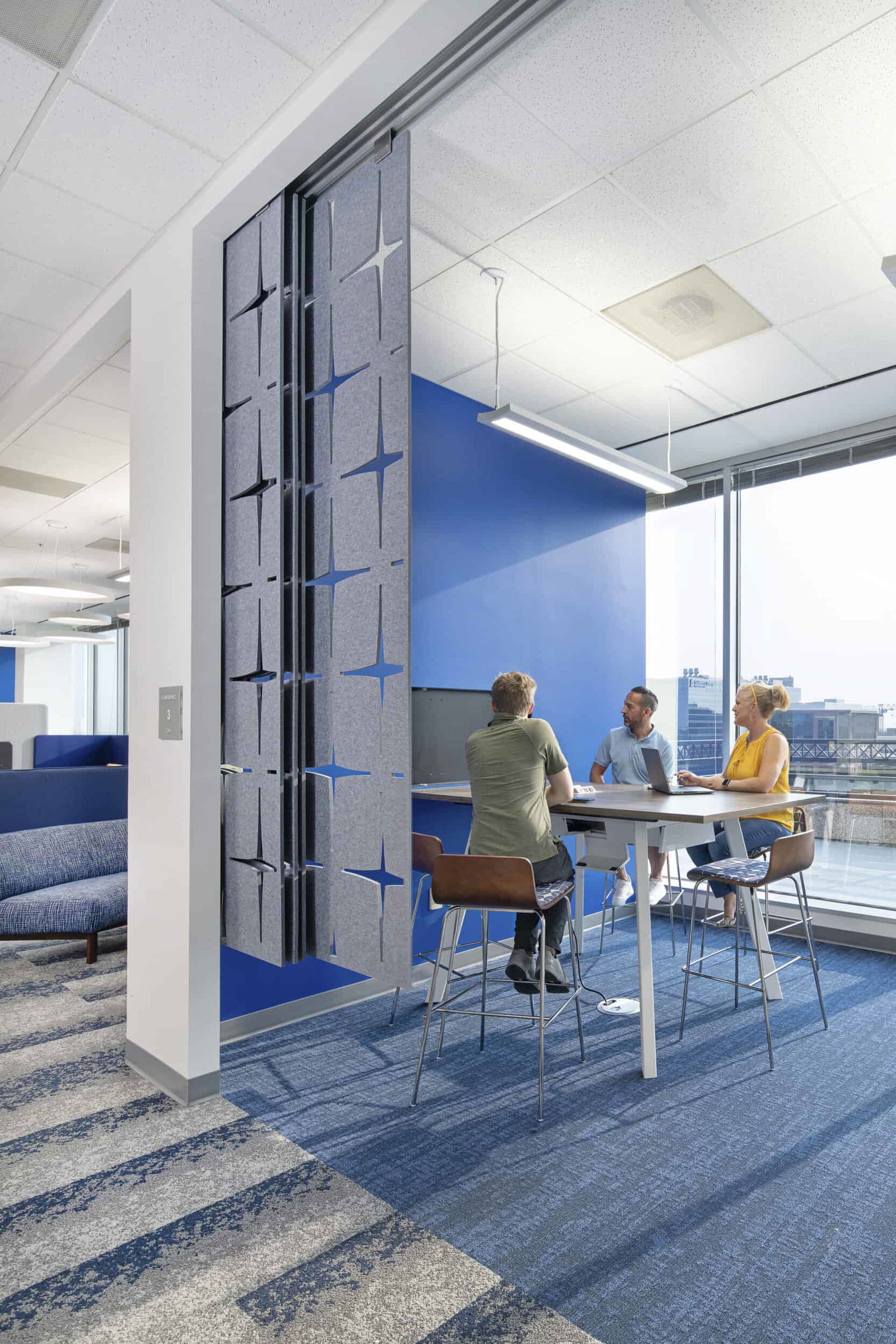 Separated from the rest of the space by a decorative gray panel with geometric cutouts that allow light to pass through is a standing-height workspace where three people sit on bar-height chairs. They are all looking at a monitor on the end of the table. Large windows provide natural light and city views throughout the space. The design showcases collaborative seating, consistent with post-pandemic flexible office environments. Blue is used as a unifying design element in wall accents, furniture, and carpeting.
