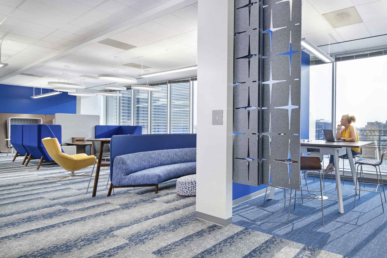 Modern office space divided into multiple work zones. On the left, a collaborative lounge area features blue high-backed booths, a blue sofa, yellow accent chair, and a patterned ottoman on blue and white patterned carpet. On the right, separated by a decorative gray panel with geometric cutouts that allow light to pass through, is a standing-height workspace where a person in a yellow top works at a laptop with bar-height chairs nearby. Large windows provide natural light and city views throughout the space. The design showcases various working styles with both collaborative seating and individual workspaces, consistent with post-pandemic flexible office environments. Blue is used as a unifying design element in wall accents, furniture, and carpeting.