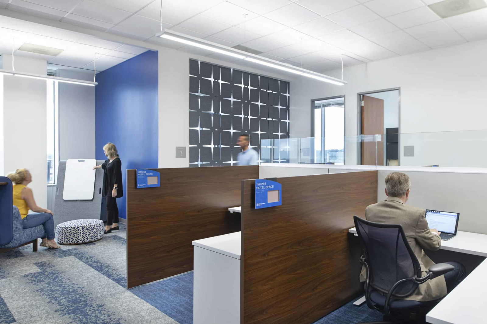 Interior of a modern office workspace featuring multiple work zones. In the foreground, individual workstations with wooden privacy panels display blue 'Hotel Space' reservation tags. A person in a beige suit works at a desk with a laptop. To the left, a casual collaboration area includes blue seating and a patterned ottoman, where two people appear to be having a discussion near a whiteboard. A blue accent wall coordinates with blue carpeting throughout. The background features a decorative black and white geometric wall panel, with a person walking past it. The space demonstrates flexible work environments with both private workstations and collaborative areas, consistent with post-pandemic hybrid work models.