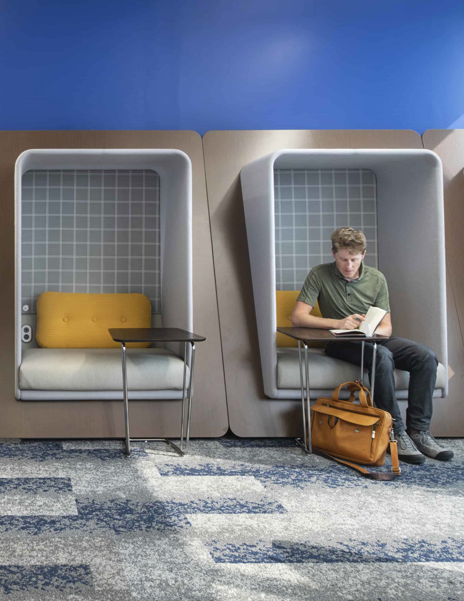 A man in a green shirt is reading a book, sitting in a pod-like enclosure with gray checkered interior backing and yellow cushioning, designed for focus work with sound absorption. The wall behind him is royal blue and matches the blue and grey patterned carpet. The design showcases different levels of privacy and work preferences in a post-pandemic flexible office environment.