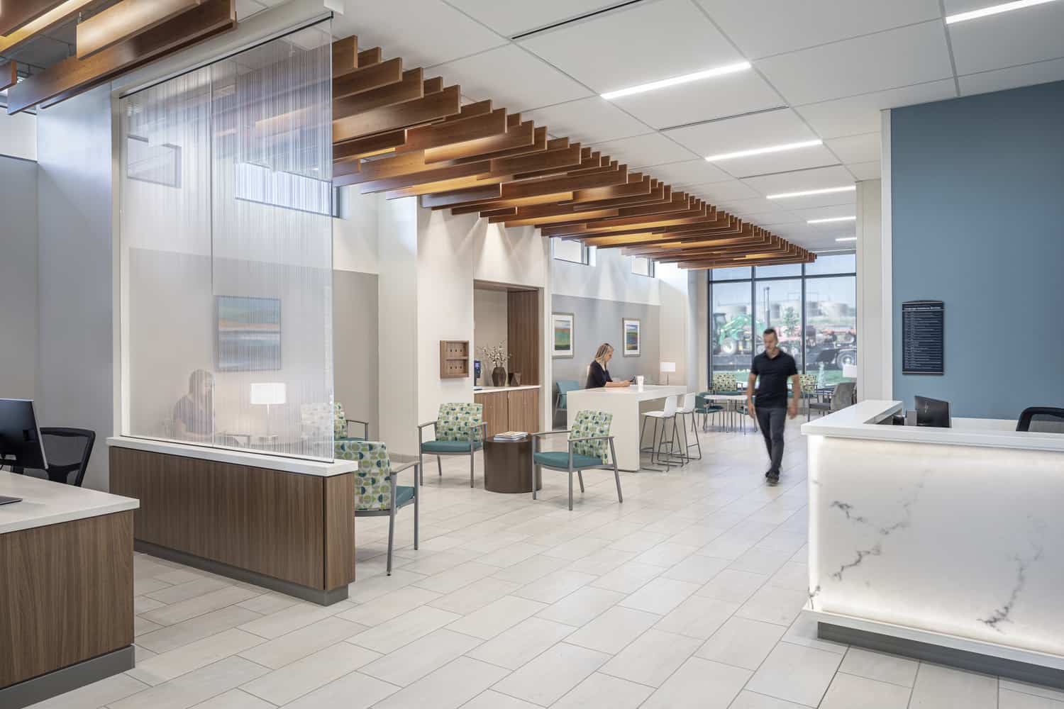 Modern waiting area within UCHealth Greeley Hospital. The space features floor-to-ceiling windows overlooking a parking lot in the very back. Blue and green armchairs and round wooden side tables are positioned near the front. A textured glass divider separates the seating area from a reception counter. Hanging wooden beams decorate the ceiling in the left side of the room, creating an illusion of a path into the waiting area.
