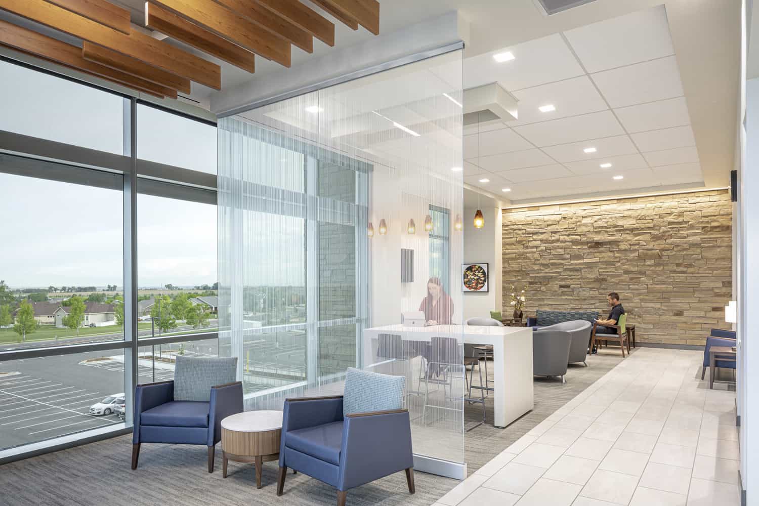 Modern waiting area within a healthcare facility. The space features floor-to-ceiling windows overlooking a parking lot and landscaped grounds. Blue armchairs and a round wooden table are positioned near the windows. A textured glass divider separates the seating area from a reception counter. The right side has a textured stone accent wall, recessed lighting, and additional seating where a person is using a laptop. Hanging wooden beams decorate the ceiling in the window area.