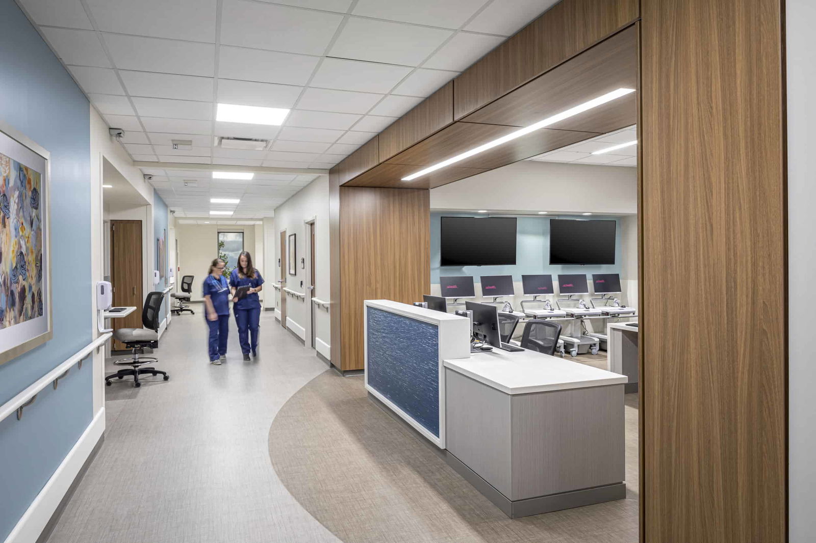 Modern hallway within the UCHealth Greeley Hospital. Two women in scrubs are walking down the hallway discussing patient information on a clipboard. On the right side, a warm wooden entryway leads to a provider work area with portable desks with monitors. There is a nurse's station in the middle of the entryway. This space was designed for with efficiency in mind for hospital staff.