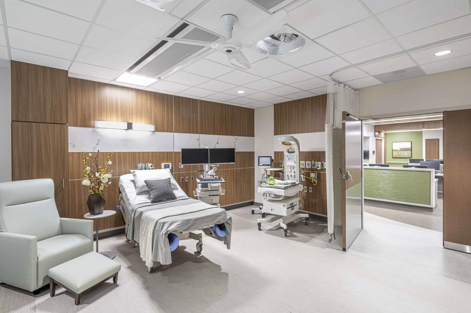 Private patient labor and delivery room within UCHealth Greeley Hospital. Warm wooden accent panels decorate the wall behind the patient bed, a typical hospital bed on wheels. A hospital bassinet for infants is beside the bed and is equipped for medical monitoring of an infant. A ceiling-mounted examination light and curtain track are visible above. A light blue recliner chair and matching footstool provide seating for visitors. Through the doorway, a green-accented nursing station is visible.