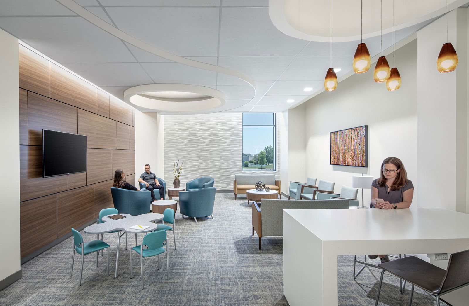 A modern healthcare waiting area with a contemporary design. The space features a mix of seating options, including blue armchairs, wooden-framed lounge chairs, and a small children's table with teal chairs. The walls are adorned with textured white paneling and warm wood accents, with a mounted flat-screen TV on one side. A large window lets in natural light, showing greenery outside. Pendant lights with amber glass shades hang from the ceiling, and recessed lighting highlights the curved ceiling design. A woman is seated at a white high-top table using her phone, while two other people are engaged in conversation in the seating area. The decor includes abstract artwork on the wall and a vase with flowers on a side table.