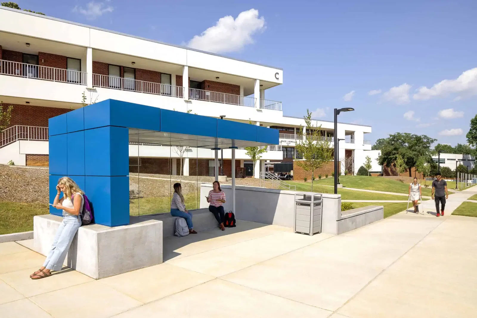 An outdoor bus shelter at Wake Tech featuring a modern design with a bright blue rectangular top structure and reflective glass panels. The shelter has concrete seating where several students are waiting - two female students are seated inside while another stands beside the structure checking her phone. In the background, a two-story white and red brick campus building is visible (marked with a 'C'), and two students can be seen walking on a concrete pathway to the right. The campus grounds include well-maintained grass areas, young trees, and landscaping. The scene is captured on a sunny day with blue sky and white clouds overhead.