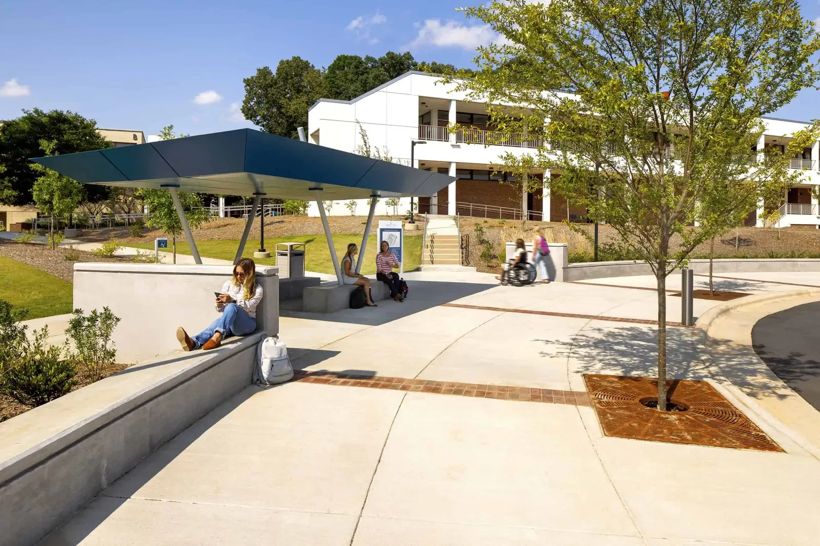 An outdoor bus shelter at Wake Tech featuring a modern design with a bright blue roof supported by diagonal cylindrical poles. The shelter has concrete seating where several students are waiting. All ramps are ADA accessible, and a student using a wheelchair is seen gliding along the sidewalk. The campus grounds include well-maintained grass areas, young trees, and landscaping. The scene is captured on a sunny day.