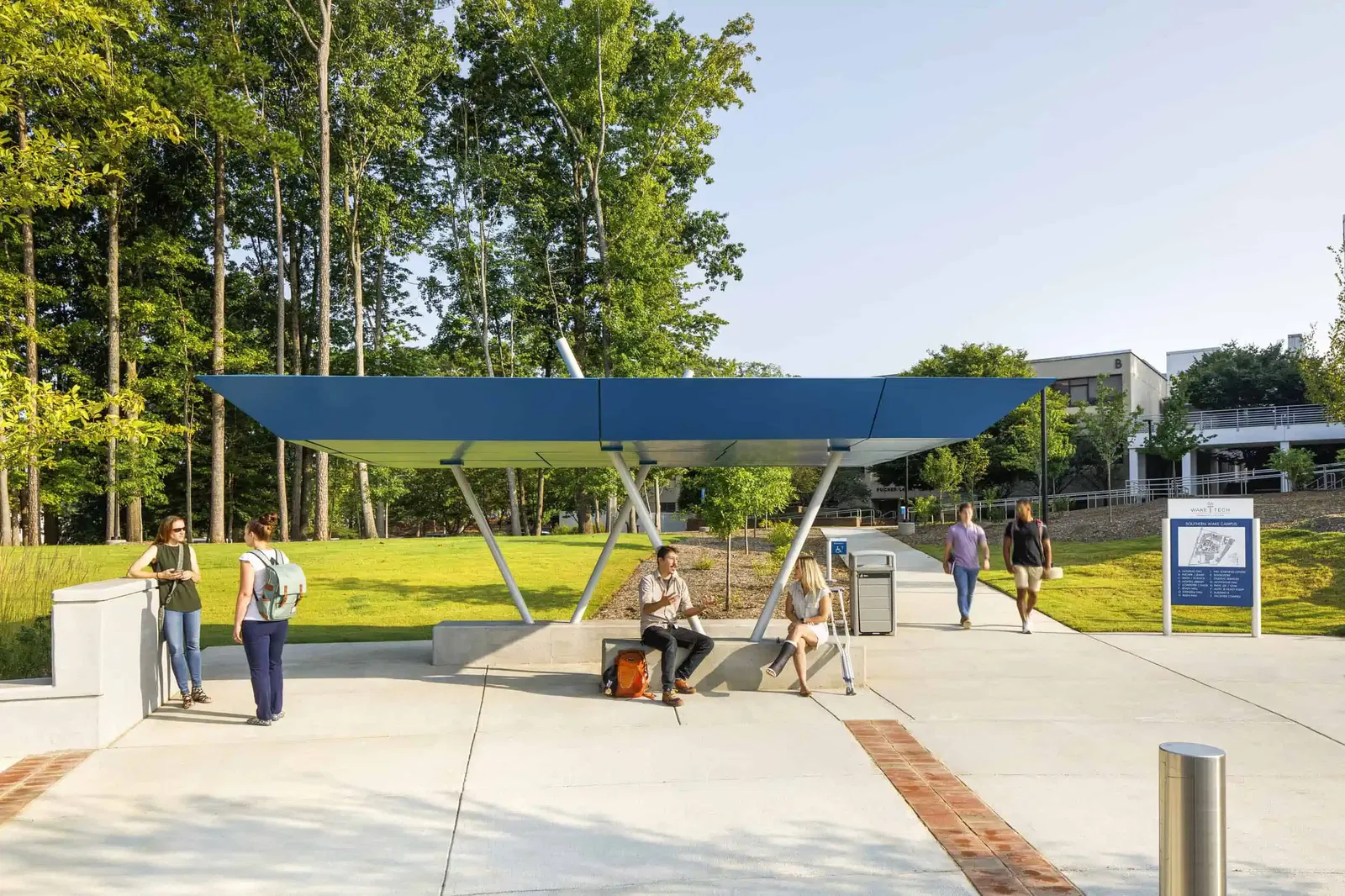 An outdoor bus shelter at Wake Tech featuring a modern design with a bright blue roof supported by diagonal cylindrical poles from the center. The shelter has concrete seating where several students are waiting. All ramps are ADA accessible. The campus grounds include well-maintained grass areas, young trees, and landscaping. The scene is captured on a sunny day.