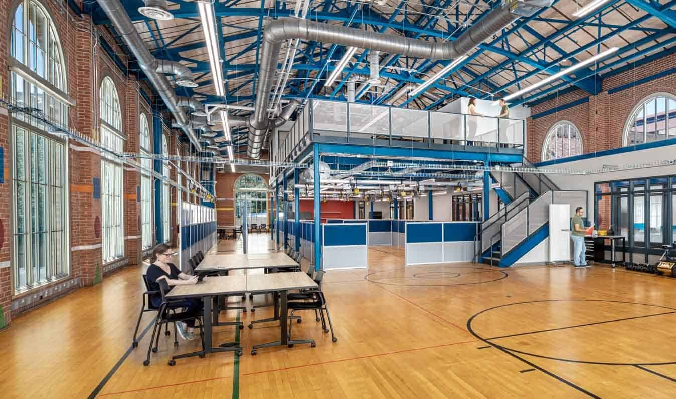 This image shows a contemporary and spacious work space within the Anna Hiss Gymnasium-turned Robotics Lab with modern workspace elements. The interior features an open ceiling with exposed silver ductwork and blue metal framing, brick walls with large arched windows, and a multi-level workspace layout. The upper level is somewhat visible though semi-transparent white guard rails. In the foreground, there is an open collaborative area with tables where a woman is sitting on her phone. The original wood floors with basketball court lines have been preserved, allowing the viewer to see that this was once a functional gymnasium.
