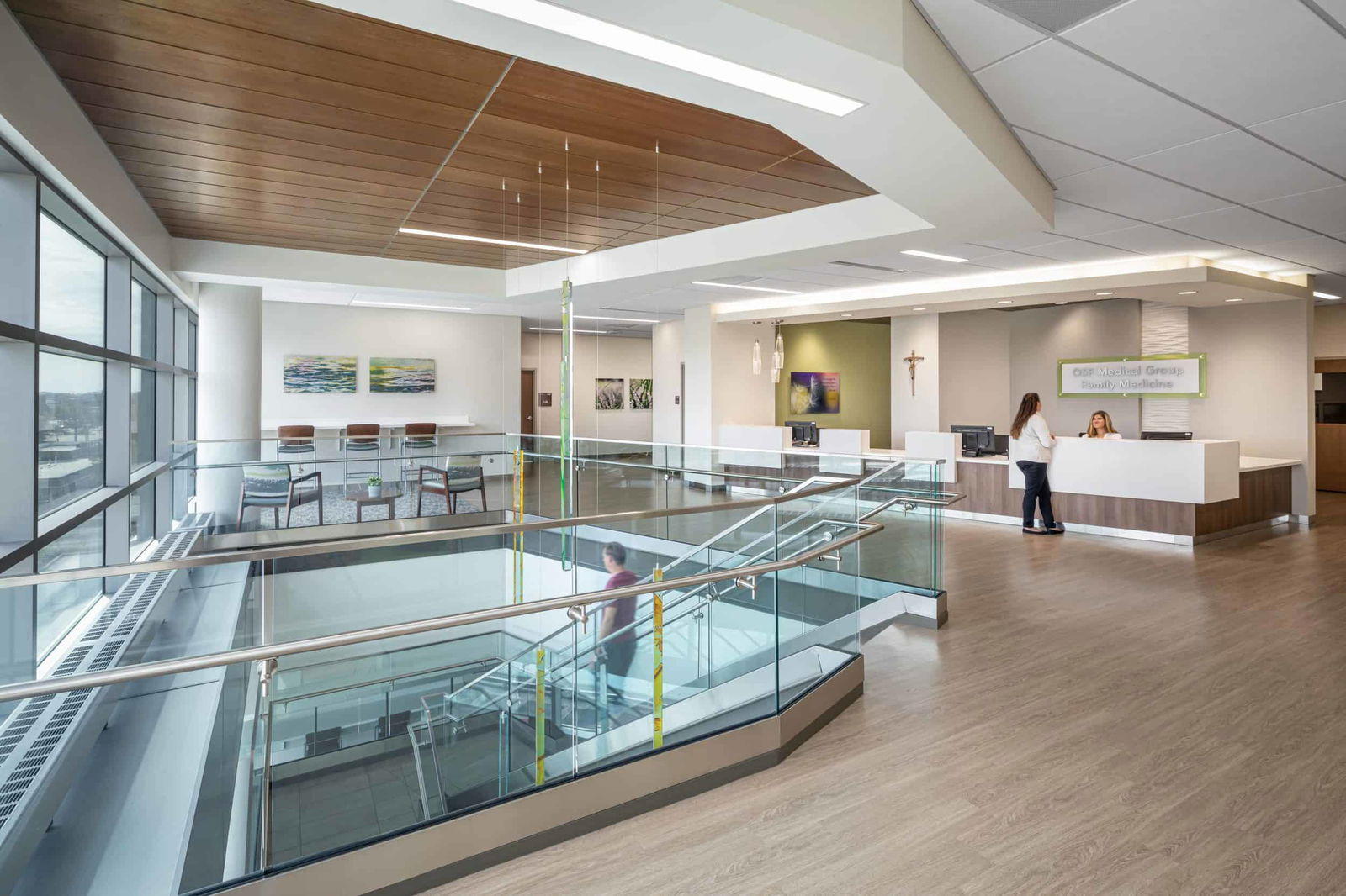 The image shows the view from the landing of the modern central staircase in the St. Joseph Medical Center. The staircase has a contemporary design with glass railings and stainless steel handrails. It features a zigzag pattern of multiple flights of stairs, creating a geometric visual effect when viewed from above. The staircase is set against wooden flooring and a wood-paneled ceiling. Hanging from the ceiling are decorative vertical elements in yellow and blue/teal colors, adding a touch of brightness to the otherwise neutral color scheme of glass, metal, and light-colored surfaces. There is also a reception desk with green accents where two women are engaged in conversation. The overall design creates an open, airy feeling with natural light flowing through the multiple levels of the building.