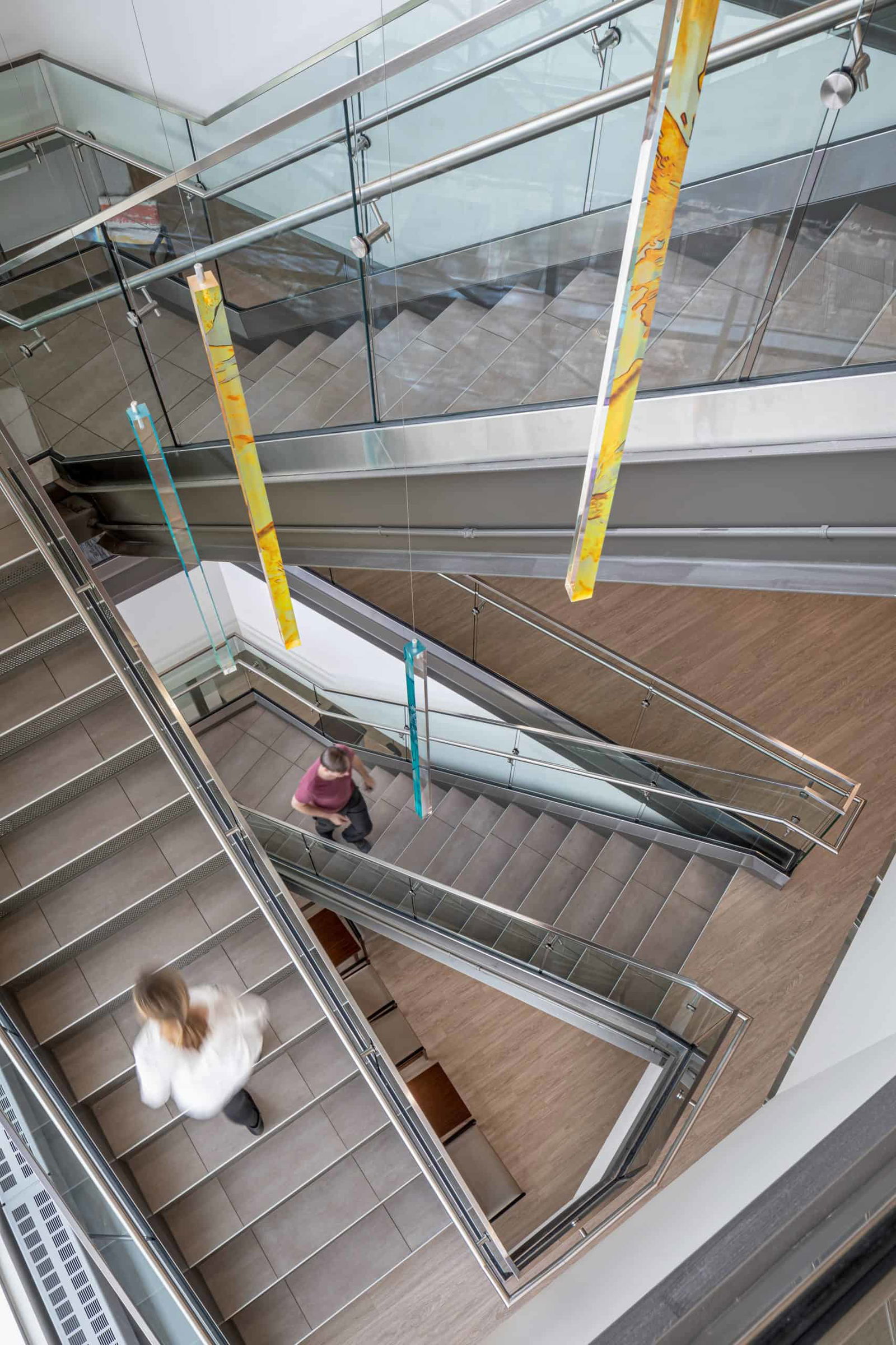 The image shows a modern staircase in the St. Joseph Medical Center. The staircase has a contemporary design with glass railings and stainless steel handrails. It features a zigzag pattern of multiple flights of stairs, creating a geometric visual effect when viewed from above. The staircase is set against wooden flooring on the landing areas, with light-colored treads on the steps themselves. Hanging from the ceiling are decorative vertical elements in yellow and blue/teal colors, adding a touch of brightness to the otherwise neutral color scheme of glass, metal, and light-colored surfaces. These colorful elements appear to be art installations that run vertically through the stairwell space. The overall design creates an open, airy feeling with natural light flowing through the multiple levels of the building.