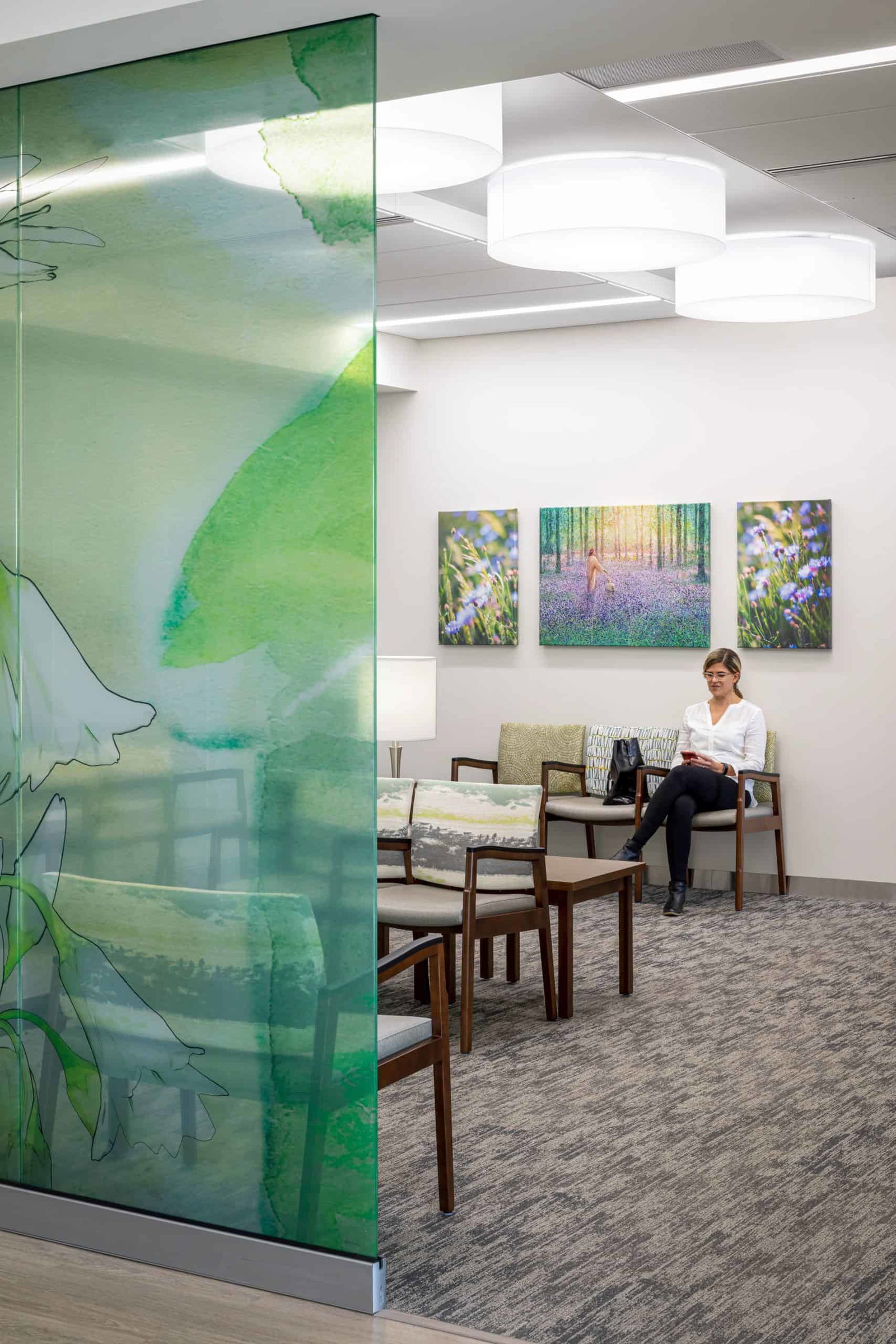 The image shows an enclosed lobby area within the St. Joseph Medical Center. On the left side, a decorative glass panel separates the waiting area from another space that cannot be seen. This glass wall features a semi-transparent nature-inspired design in blue and green tones, possibly depicting plants. Beyond the panel, there is a seating area of green and grey fabric chairs with wooden bases arranged in U shapes. A woman is sitting in a chair and is scrolling on her phone. The overall design is clean, bright, and welcoming with plenty of natural light, creating a calming atmosphere typical of contemporary healthcare environments.