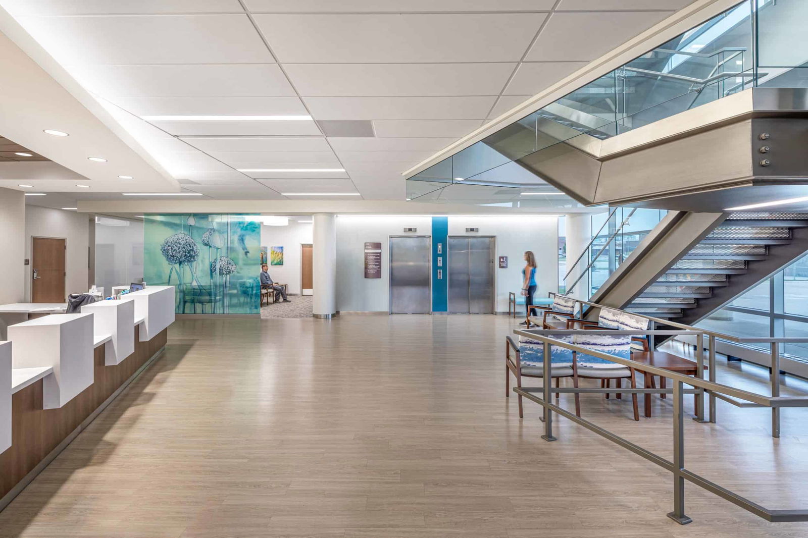 The image shows a lobby area within the St. Joseph Medical Center. On the left side, there's a long reception desk with a white counter and wooden base. The open, spacious lobby features light wood-look flooring and a white drop ceiling with recessed lighting. In the center back of the image, there are two elevator doors with a blue accent panel between them. A person in light blue clothing can be seen standing near the elevators. To the right side of the image, there's a modern staircase with glass and metal railings leading to an upper level. Near the staircase, there's a small seating area with a few chairs. The left wall features a decorative glass panel with a nature-inspired design in blue and green tones, possibly depicting dandelions or similar plants. The overall design is clean, bright, and welcoming with plenty of natural light, creating a calming atmosphere typical of contemporary healthcare environments.