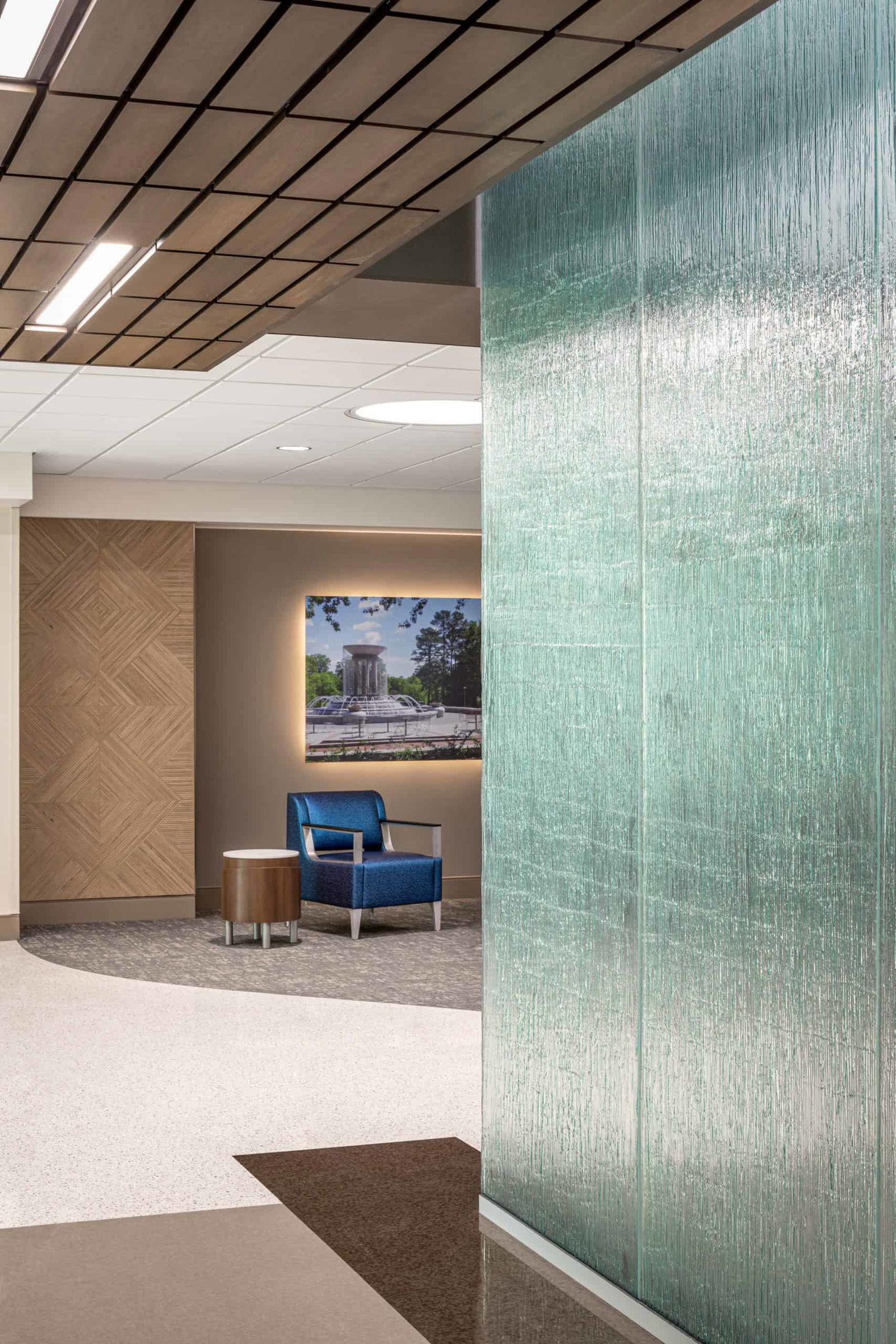 The image shows a close-up view of the design elements in a reception area in the WakeMed Cary Hospital. Suspended wood ceilings with lights, a textured glass wall, decorative wood accents on walls, and modern seating area with high-end blue furniture contribute to a modern feel. The room has clean, modern design with neutral colors and a large picture of a fountain with lighting behind it, giving it a glowing illusion.