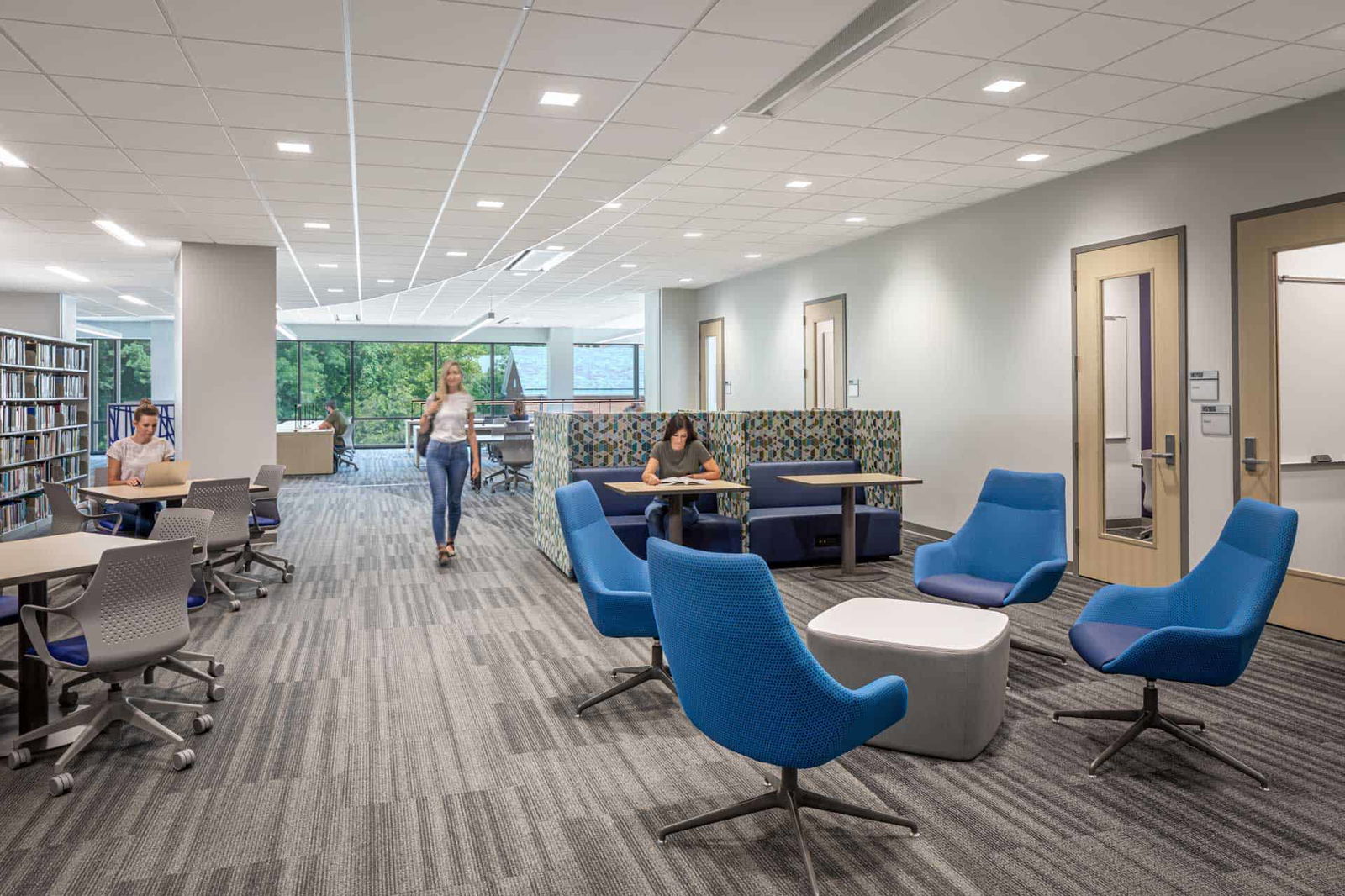 This image shows a modern academic space within the Butler University Sciences Complex with multiple functional areas. On the left side are two sets of tables, each with four blue and grey office chairs. Behind these tables are library-style bookshelves. On the right side are sets of modern blue accent chairs situated around a blue and grey ottoman. The room has a dropped ceiling with recessed lighting and gray carpeted flooring throughout. The overall design combines open collaborative spaces with more private work areas in a contemporary academic environment.