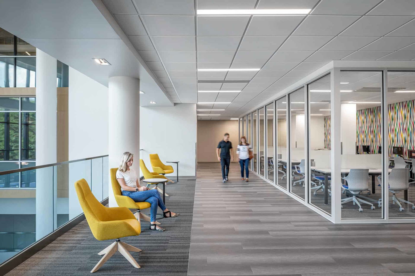 This image shows a modern academic space within the Butler University Sciences Complex with multiple functional areas. On the right side is an enclosed classroom with glass walls, making the interior of the room visible. Beyond the glass walls of the classroom is a multi-colored vertically textured mural in shades of yellow, red, green, and blue. The classroom has rows of white tables with two grey and blue office chairs at each table. Outside on the left side of the photo are two sets of modern, bright yellow chairs. The center is a hallway with laminate grey flooring. The space has a dropped ceiling with recessed lighting. The overall design combines open collaborative spaces with classrooms in a contemporary academic environment.