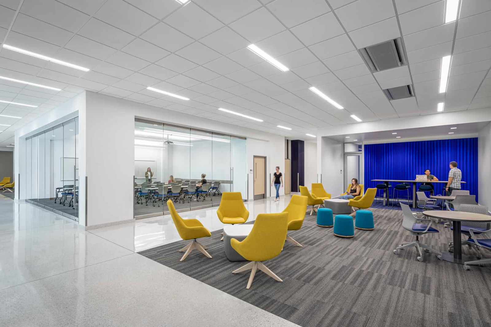 This image shows a modern academic space within the Butler University Sciences Complex with multiple functional areas. In the background is an enclosed classroom with glass walls, so the interior is visible from the common area. Closer to the camera is a gathering space with modern, bright yellow chairs, teal ottomans, circular grey tables with three glue and grey office style chairs against a dramatic royal blue wall. The space features distinctive flooring types signifying the boundaries of the different areas. The space has a dropped ceiling with recessed lighting. The overall design combines open collaborative spaces with classrooms in a contemporary academic environment.