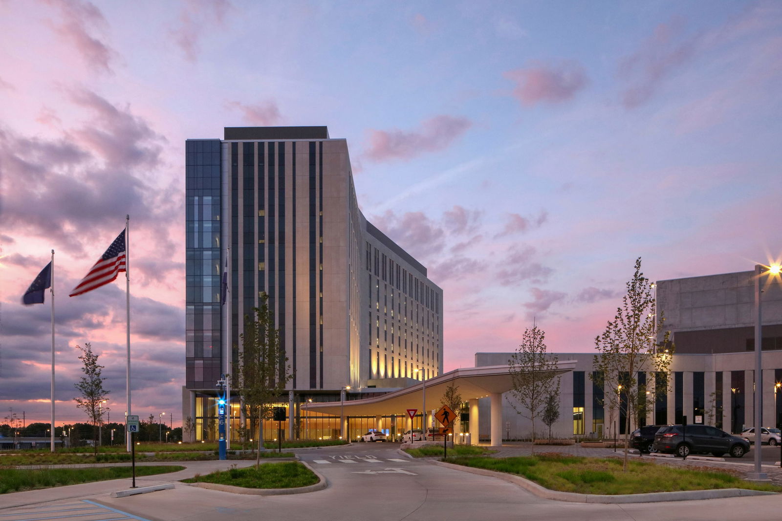 This photo showcases Marion County Community Justice Campus in Indianapolis at dusk with a dramatic pink, purple, and blue sky. The complex features a striking combination of modern architectural elements, including a tall glass-and-concrete tower and a sprawling, lower-rise structure behind it. A driveway leads to a covered drop-off area at the main entrance, flanked by landscaped plantings and multiple flagpoles. The design blends clean lines, vertical window elements, and open green spaces, emphasizing both functionality and a welcoming civic presence. Warm lighting is coming through the windows, giving the building a beautiful glow.