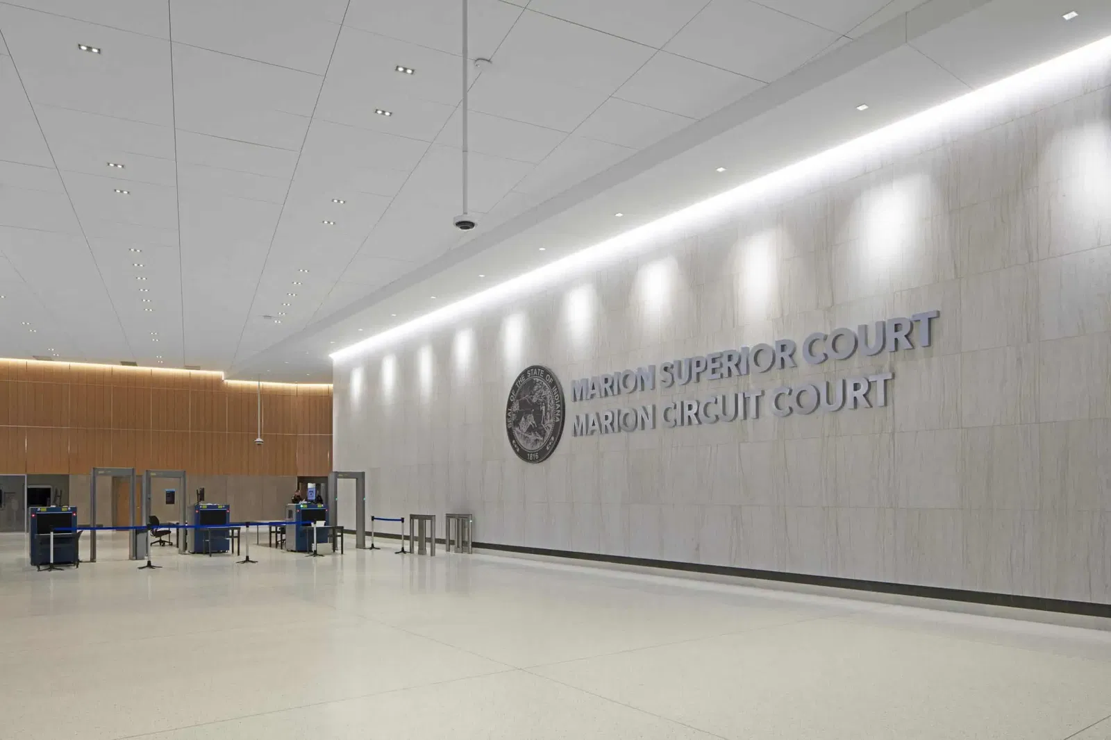 The image shows the interior lobby of the Marion Superior Court and Marion Circuit Court. The space features a minimalist, modern design with a large white marble or stone wall that prominently displays the court names in silver metallic lettering alongside what appears to be an official seal or emblem. The ceiling is white with recessed lighting, and there's elegant ambient lighting along the wall. To the left side of the image, there's a security checkpoint area with metal detectors and screening stations, typical of courthouse entry procedures. The floor is polished light-colored stone or tile, and the overall aesthetic is clean, spacious, and formal, befitting a judicial building. The design combines functionality with a sense of dignity appropriate for a courthouse.