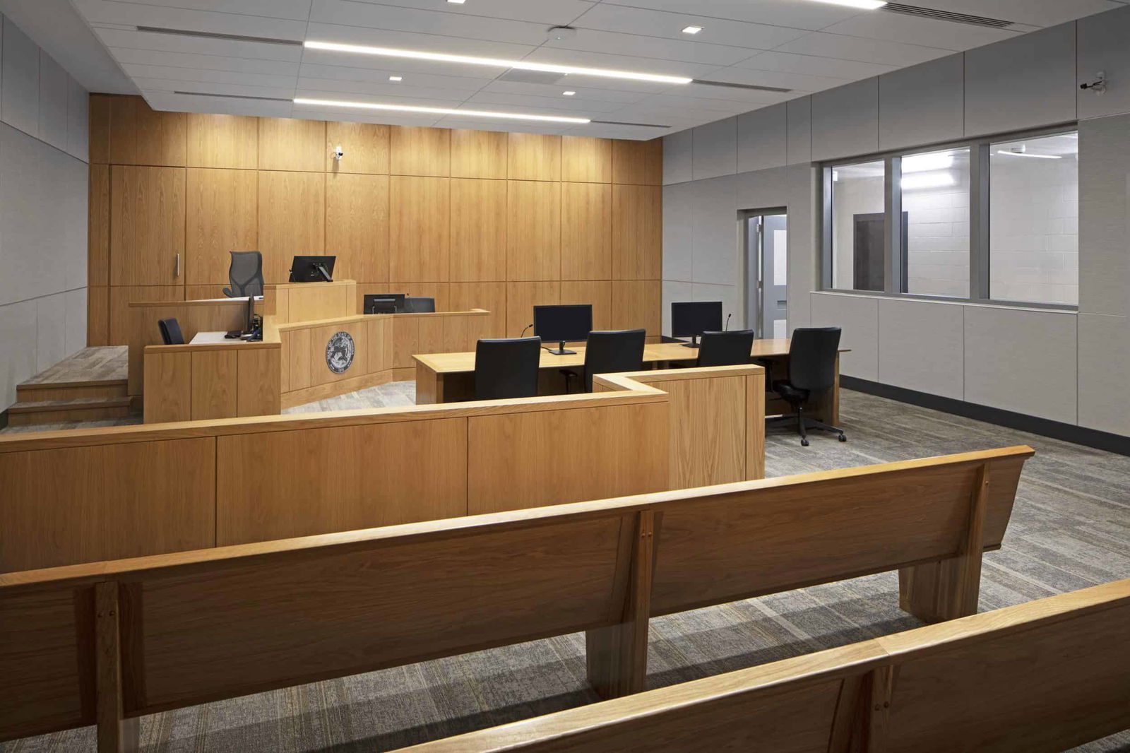 The image shows a modern Marion County courtroom interior. It features a clean, contemporary design with warm wood paneling on the judge's bench and surrounding walls. The judge's area is elevated and includes a desk with what appears to be a small emblem or seal embedded in the front. There are several computer monitors visible on the bench and at attorney tables. In the foreground, wooden spectator benches are arranged in rows, facing the front of the courtroom. The attorneys' tables are positioned between the gallery and the judge's bench, equipped with chairs and computers. The courtroom has a neutral color palette with light wood tones and gray walls. The space is well-lit with recessed ceiling lights and has a window or viewing area on the right side wall, likely for observation or recording purposes. The overall design balances traditional courtroom formality with contemporary architectural elements, creating a professional judicial environment.