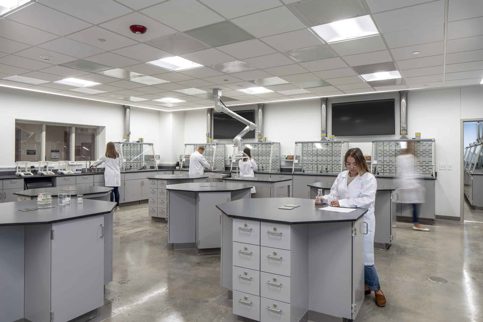 Modern laboratory with bright overhead lighting and neutral gray tones. Several scientists in white lab coats are working at large hexagonal lab tables equipped with storage drawers. The lab features wall-mounted storage bins, a fume hood, and an articulating extraction arm suspended from the ceiling. The floor is polished concrete, and the overall atmosphere is clean and organized.