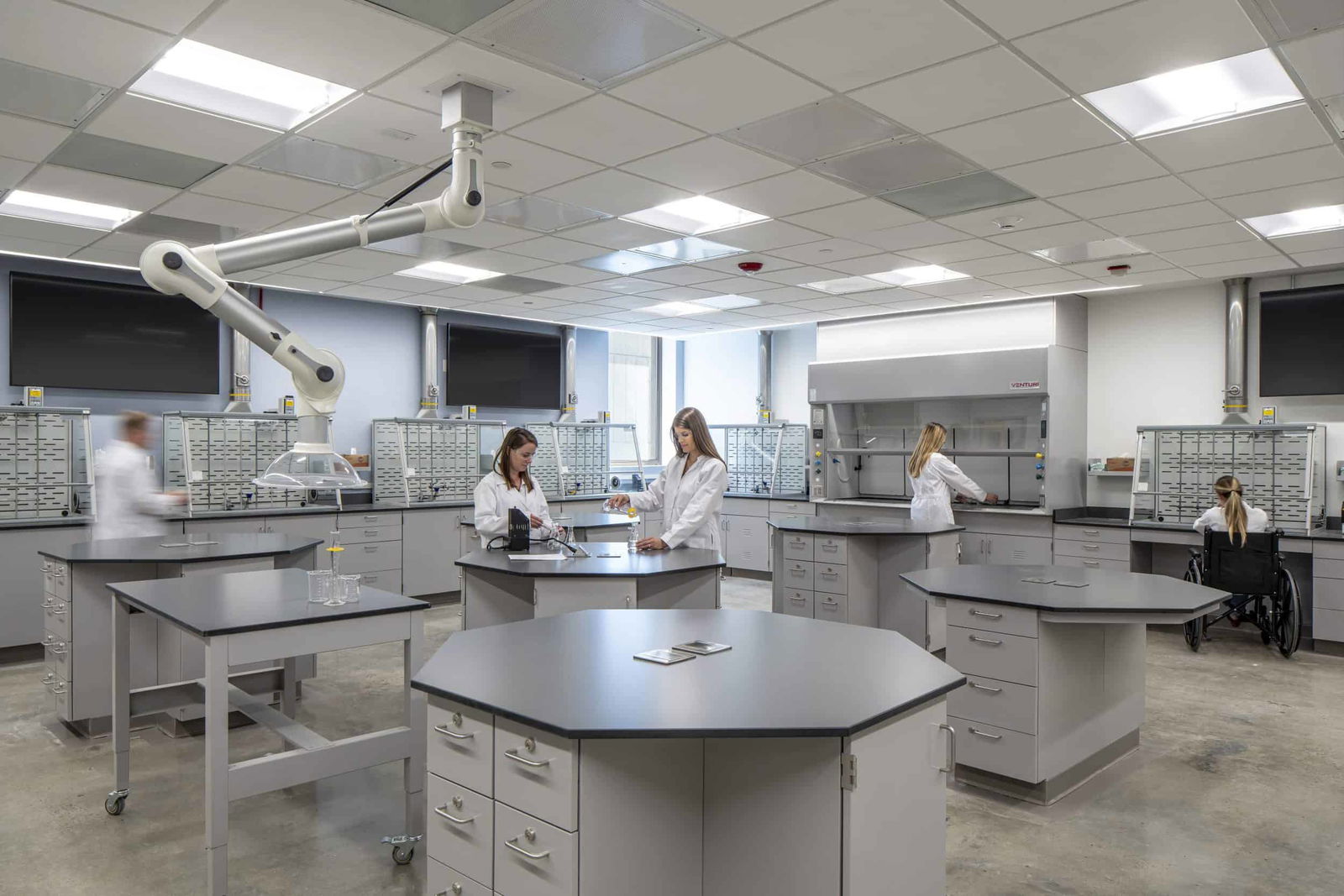 Modern laboratory with bright overhead lighting and neutral gray tones. Several scientists in white lab coats are working at large hexagonal lab tables equipped with storage drawers. One person in a wheelchair is working at a station, highlighting accessibility. The lab features wall-mounted storage bins, a fume hood, and an articulating extraction arm suspended from the ceiling. The floor is polished concrete, and the overall atmosphere is clean, organized, and inclusive.