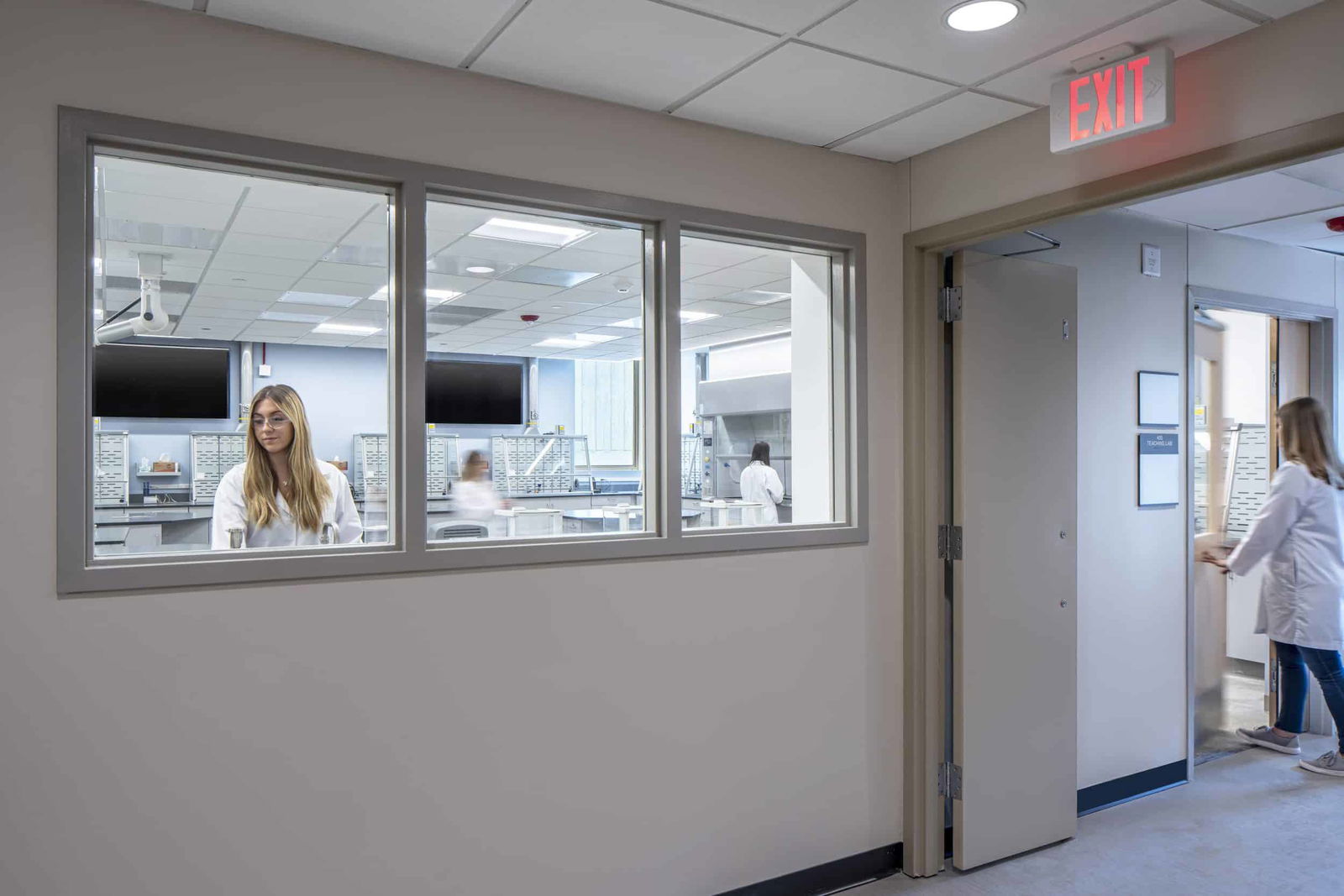 This image shows the entrance to a modern laboratory setting from a grey hallway. Windows into the laboratory show a woman in a white lab coat. The lab features white walls, a drop ceiling with integrated lighting, and extensive cabinetry with multiple small drawers along the back wall, likely for storing supplies or specimens.