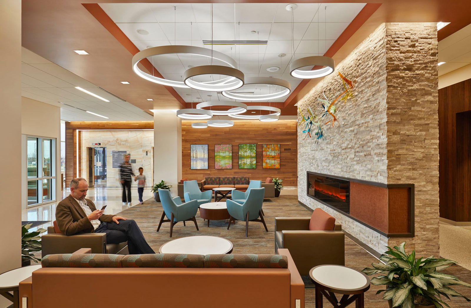 This image shows a modern healthcare facility lobby or waiting area with upscale design elements. The space features a stone accent wall with a built-in linear fireplace on the right side. The ceiling has distinctive circular pendant light fixtures arranged in an artistic pattern with warm copper/orange ceiling accents. The seating area includes several conversation groupings with light blue armchairs and brown/tan sofas arranged around circular coffee tables. A person in a brown jacket sits in one of the lounge chairs looking at a mobile device. In the background, there appears to be movement of people walking through the space. The interior design combines natural elements like wood paneling (visible on the back wall with four seasonal landscape artworks) and stacked stone with contemporary furnishings. The color palette features earthy tones with blue accents. Potted plants are placed throughout the space to add greenery. The overall atmosphere suggests a high-end medical facility or healthcare center designed to feel welcoming and less institutional than traditional clinical environments.