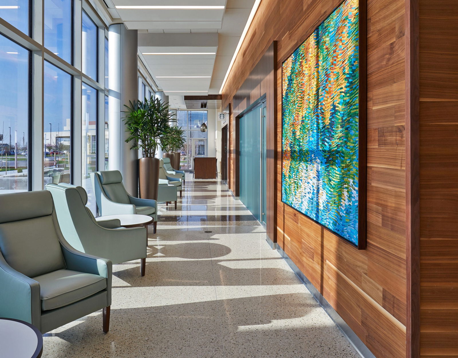 This image shows a modern healthcare facility corridor or hallway with large floor-to-ceiling windows along the left side that allow natural light to flood the space. The right wall features warm wood paneling with a vibrant, large-scale artwork in blues, greens, and gold/orange tones that creates a focal point. The corridor has a polished terrazzo or stone floor with an interesting pattern of light and shadow created by the sunlight coming through the windows. Several light blue/sage green lounge chairs are positioned along the left side, creating small waiting areas. Tall planters with indoor trees are strategically placed to bring natural elements into the space. There appears to be a doorway or entrance visible further down the corridor, possibly to a treatment room or office. The overall design combines natural materials, art, comfortable seating, and abundant natural light to create a healing environment that feels more like a high-end hotel than a clinical setting. This approach to healthcare design aims to reduce stress and create a more welcoming experience for patients and visitors.