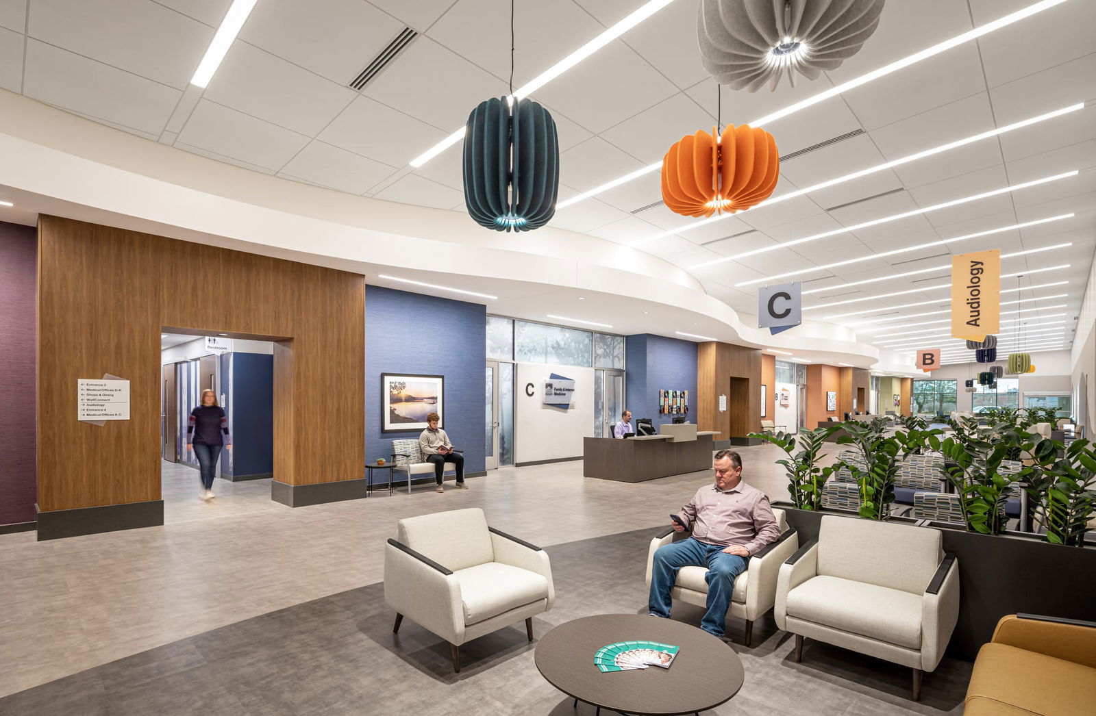 Interior of a converted mall healthcare complex, showing another section of the spacious lobby. Colorful pendant lights in teal and orange hang from white linear ceiling lights. Wayfinding signs marked 'C' and 'Audiology' are visible. Modern seating areas with white chairs and potted plants are arranged near wood-paneled walls and blue-painted sections. A person sits in one of the chairs, and another walks through a doorway in the background.