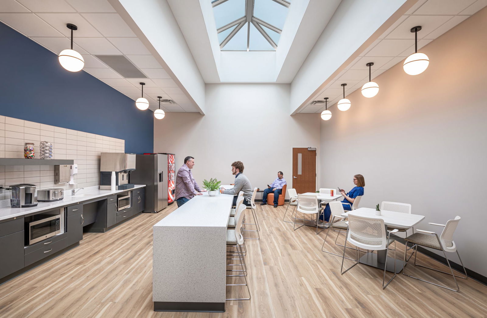 Modern break room or staff lounge within the converted mall healthcare facility. A large skylight illuminates the room with hanging globe pendant lights. A long white kitchen island with bar stools is in the foreground, with kitchen appliances and a blue accent wall to the left. Several staff members are seated at white tables and chairs, some conversing and others using electronic devices. The room has light wood-tone flooring and a clean, contemporary design.