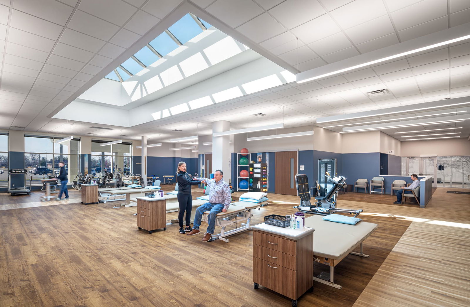 Physical therapy or rehabilitation room within the converted mall healthcare facility. A large geometric skylight floods the spacious room with natural light. Examination tables, medical equipment, and wooden cabinets are arranged throughout the space. A healthcare professional is talking with a patient seated on an examination table. The room has wood-tone flooring, white walls, and blue accent panels. Medical exercise equipment and other treatment stations are visible in the background.