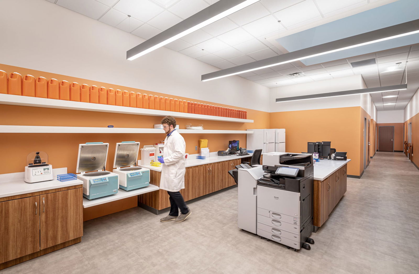 Laboratory workspace with a technician in a white lab coat. The room has orange and white walls, wood-tone cabinetry, and linear ceiling lights. Multiple scientific equipment pieces are visible, including refrigerated units and a large printer/copier. Rows of orange storage containers are neatly arranged on shelves along the wall. The space has a clean, organized medical research or diagnostic lab appearance.