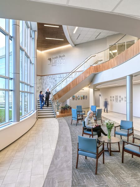 SBL Bonutti Clinic Interior Lobby with with a large wood, glass, and metal staircase leading to the second floor which has a curved loft. Large floor-to-ceiling windows allow the spacious atrium to be flooded with light. This image showcases BSA's expertise in interior design.