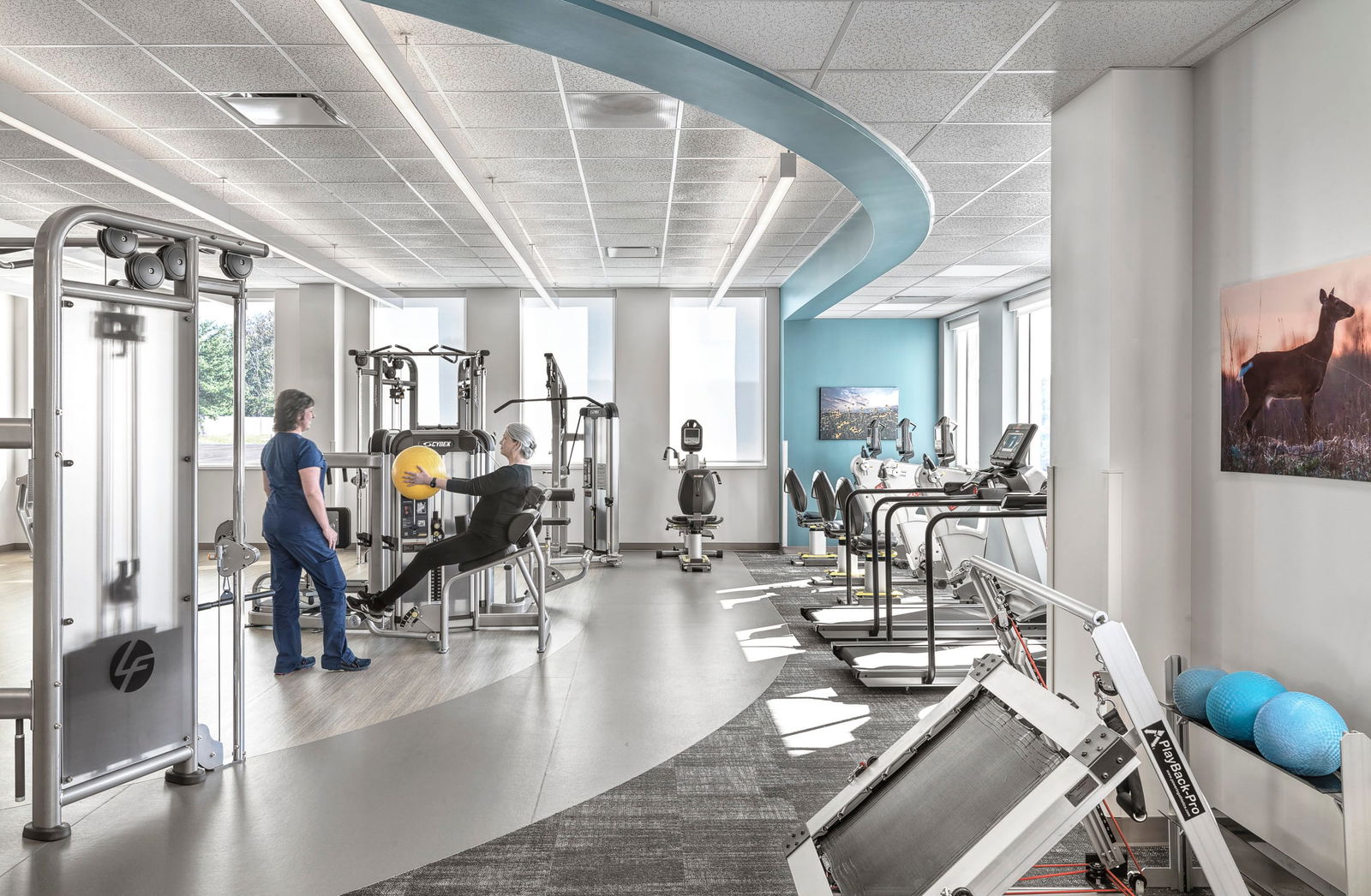 SBL Bonutti Clinic Interior Lobby patient gym featuring modern, curved ceiling designs. A patient is using an exercise machine with the help of a staff member in navy scrubs. Exercise equipment fills the room. This image showcases BSA's expertise in designing spaces that prioritize patient care.