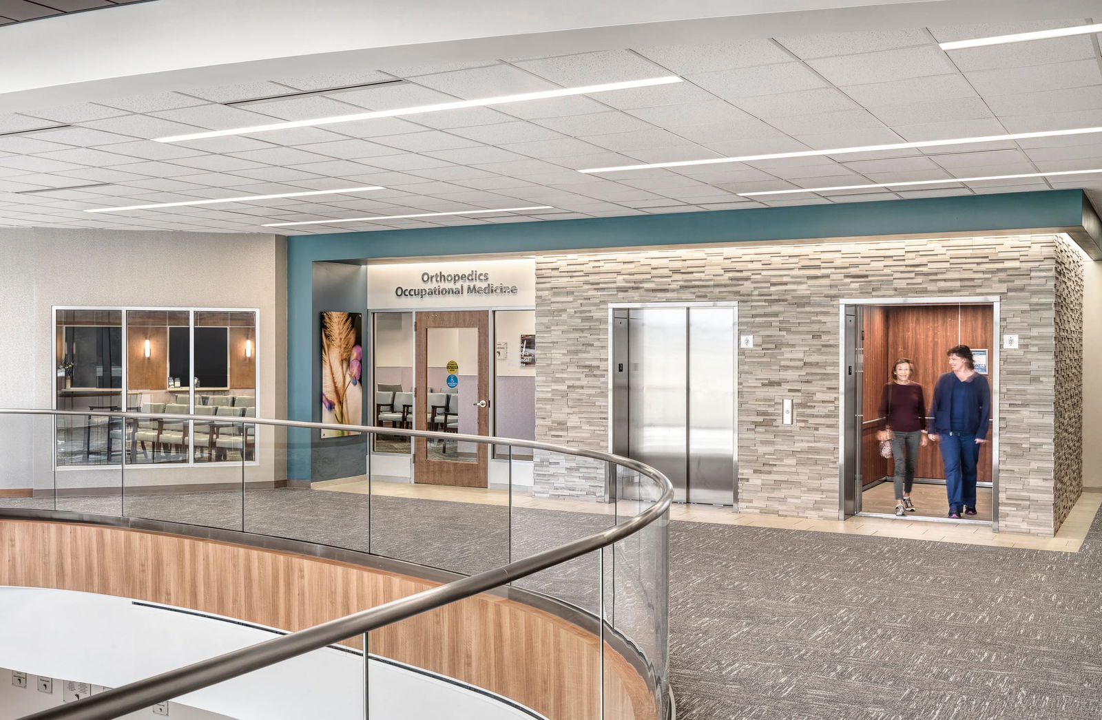 SBL Bonutti Clinic interior second floor lobby with two elevators and an entrance to the Orthopedics Occupational Medicine lobby. Glass railings and a curved loft-like element allow the first floor to be visible. Two people are getting off the elevator. This image showcases BSA's expertise in interior design.