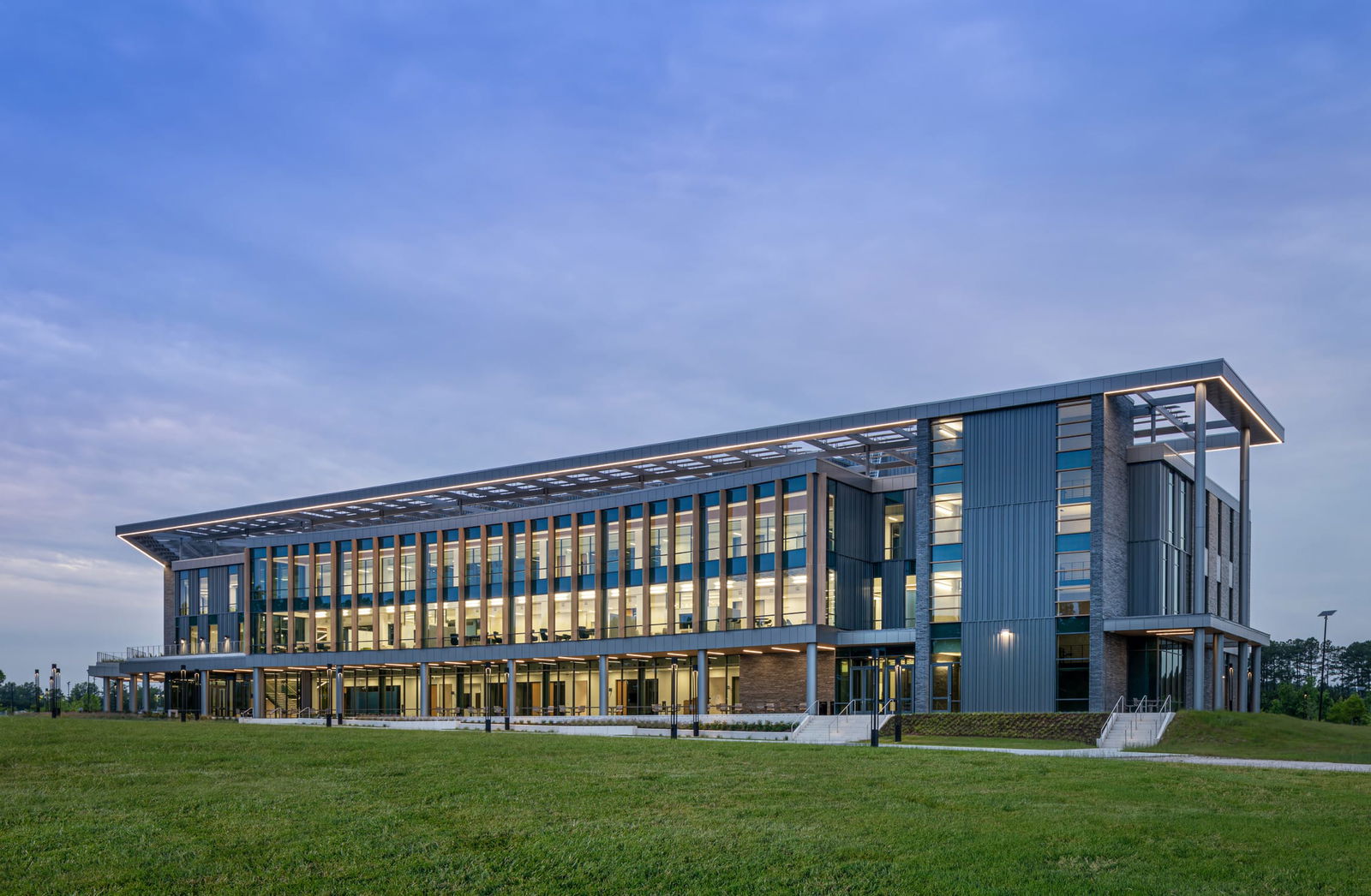 Exterior view of the Wake Tech Education and Innovation Center at dusk, designed with clean lines, expansive glass, and a prominent overhanging roof. The multi-story building features vertical fins, modern metal and brick finishes, and warm interior lighting that glows through the large windows, all set against a backdrop of open green space and a soft evening sky.