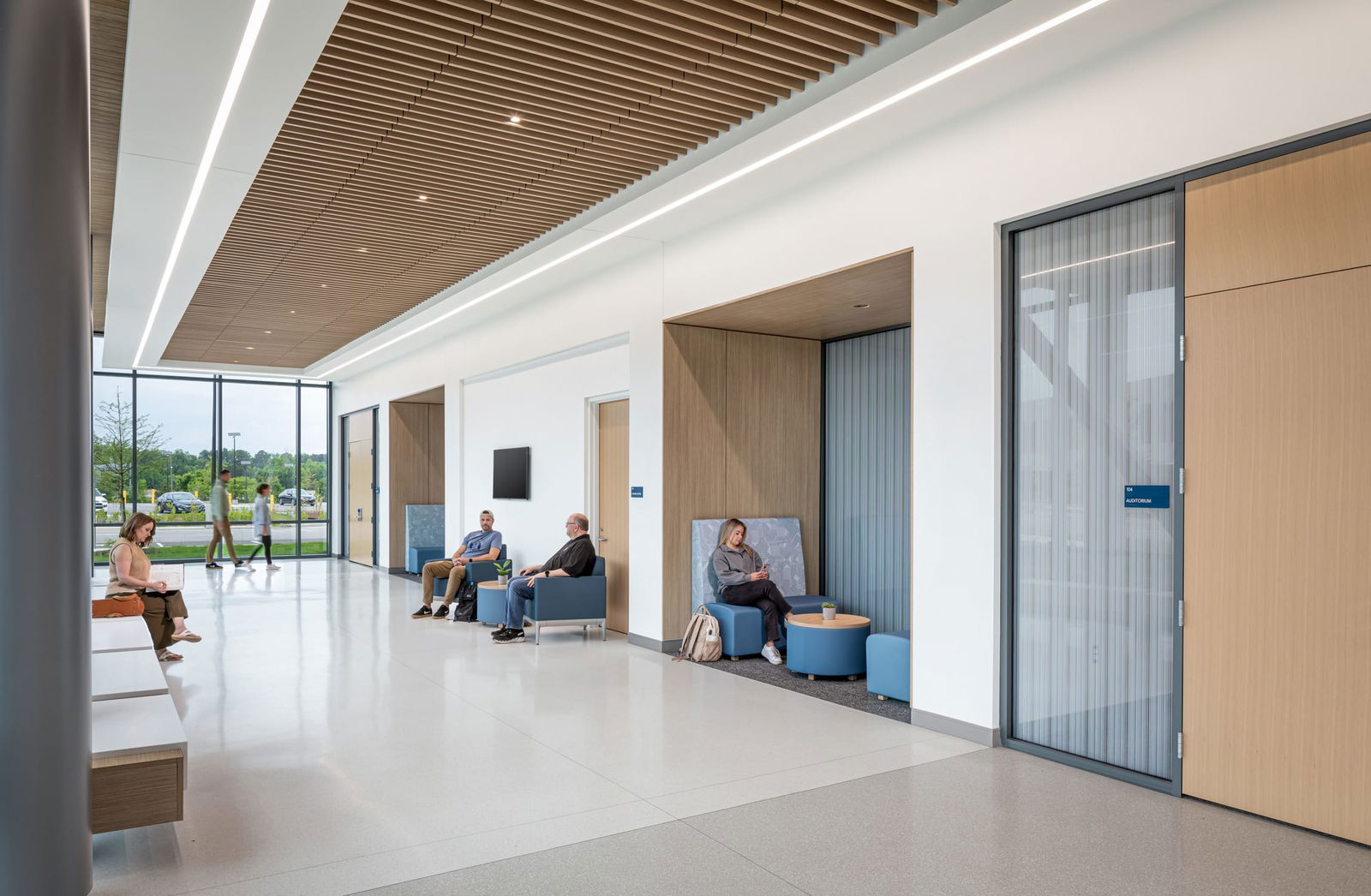 Interior view of a bright, modern student commons area at the Wake Tech Education and Innovation Center. The space features floor-to-ceiling windows with views of the green landscape outside. A slatted wood ceiling detail with recessed lighting adds warmth and texture along the central spine of the space. Students are seen studying in upholstered booths, sitting on ottomans, and walking through space, creating a lively and collaborative atmosphere.