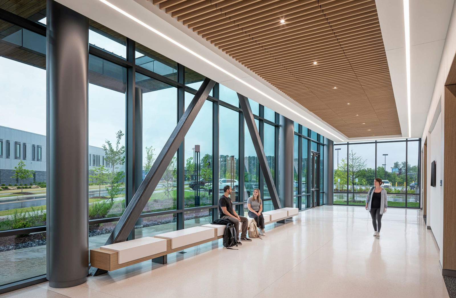 Interior view of a bright, modern student commons area at the Wake Tech Education and Innovation Center. The space features floor-to-ceiling windows with metal beams in horizontal, vertical, and diagonal patterns. Overhead, a slatted wood ceiling detail with recessed lighting adds warmth and texture along the central spine of the space. Students are seen chatting on a long bench that spans the length of the windows.