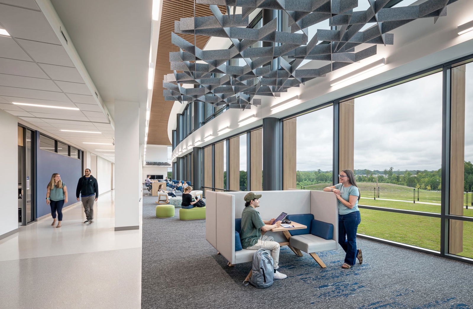 Interior view of a bright, modern student commons area at the Wake Tech Education and Innovation Center. The space features floor-to-ceiling windows with views of the green landscape outside. Overhead, an array of sculptural, geometric acoustic baffles hangs from the ceiling in dynamic, crisscrossed patterns, paired with linear pendant lighting. A slatted wood ceiling detail adds warmth and texture along the central spine of the space. Students are seen studying in upholstered booths, sitting on ottomans, and walking through the adjacent corridor, creating a lively and collaborative atmosphere.