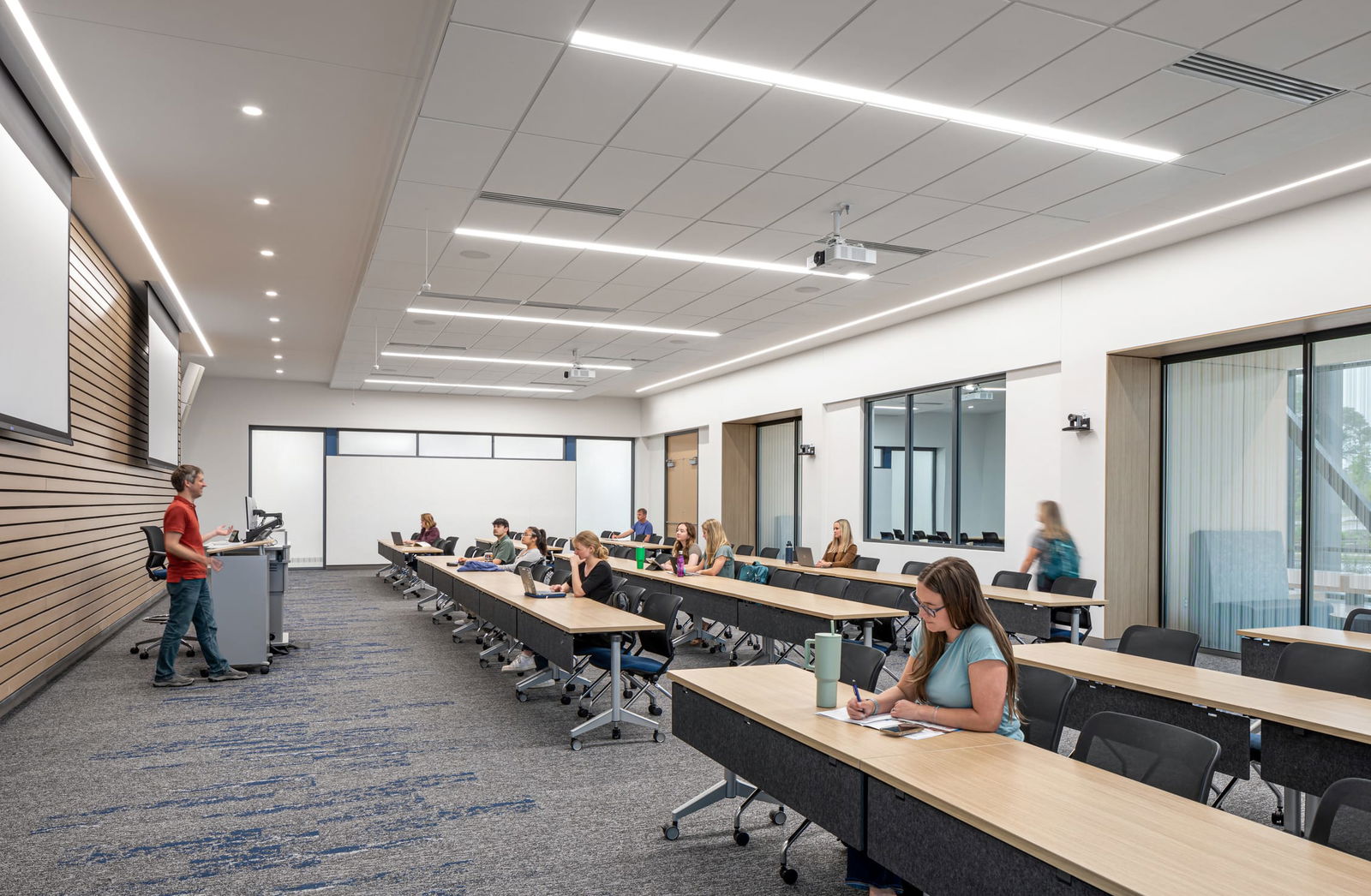 A modern classroom at Wake Tech Community College featuring tiered rows of light wood tables with black chairs. A male instructor in a red shirt stands at a podium on the left side of the room while students are seated throughout the classroom. The space has contemporary design elements including recessed lighting in the white ceiling, carpeted flooring with blue patterns, and wooden accent panels on the left wall. Ceiling-mounted projectors, a whiteboard, and internal windows looking into adjacent rooms complete the educational environment. Students are engaged in note-taking and classroom activities.