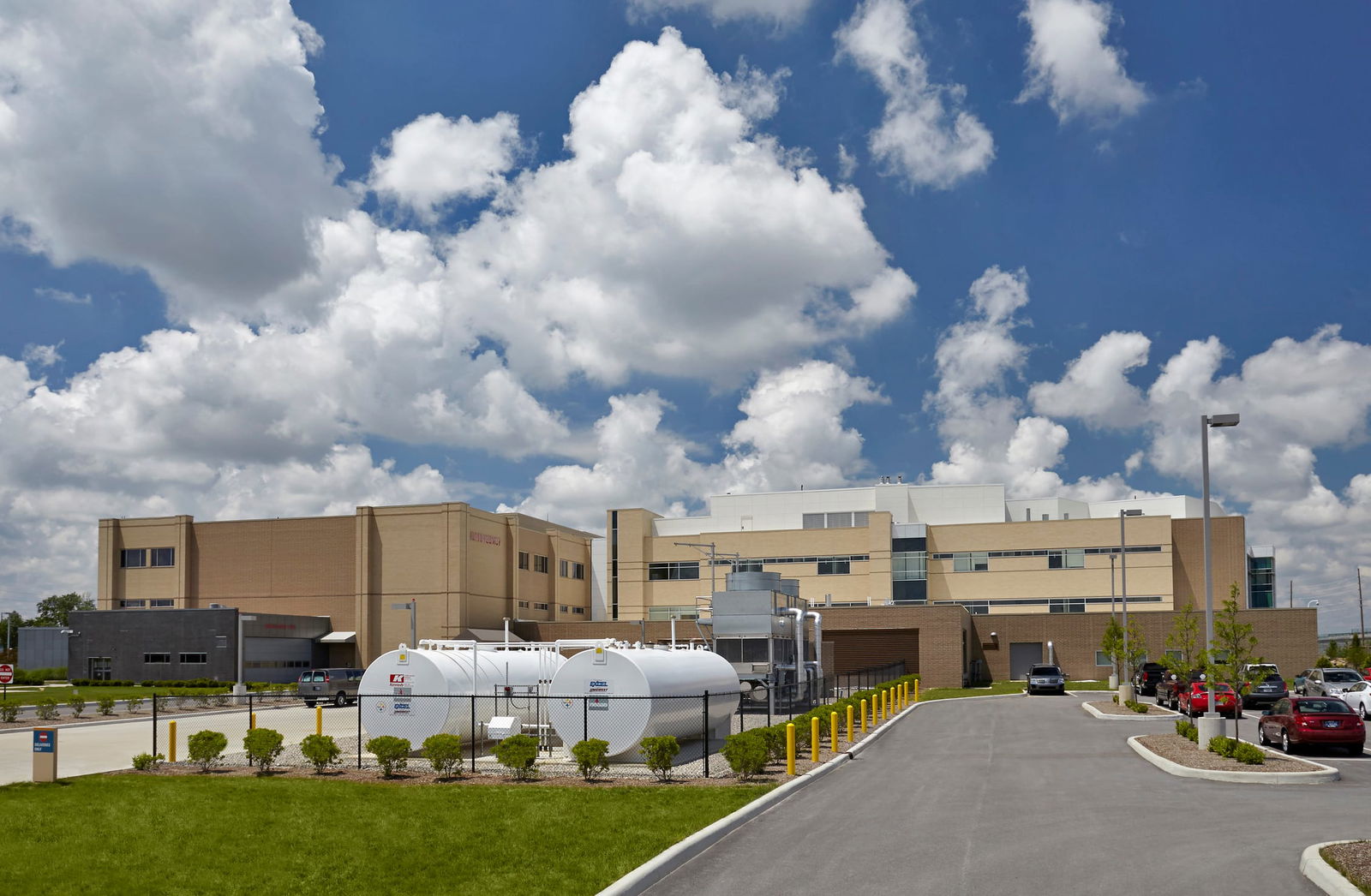 The large, multi-story Ascension St. Vincent Fishers Hospital with a beige brick exterior and modern white additions on the upper floors. The foreground features a fenced-off utility area with large white fuel tanks, industrial pipes, and a small landscaped section with shrubs. The purpose of this image is to convey BSA's engineering expertise within this project, highlighted by the utility area.