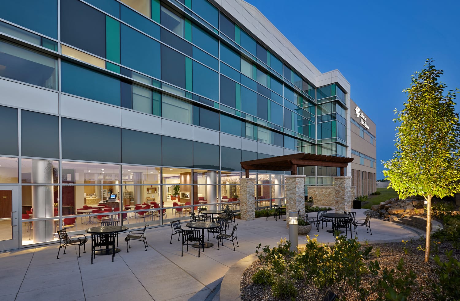 Glass-paneled exterior of Ascension St. Vincent Fishers Hospital, featuring blue and green tinted windows and a concrete patio with tables and chairs with landscaping. This image show's BSA's expertise in exterior architecture.