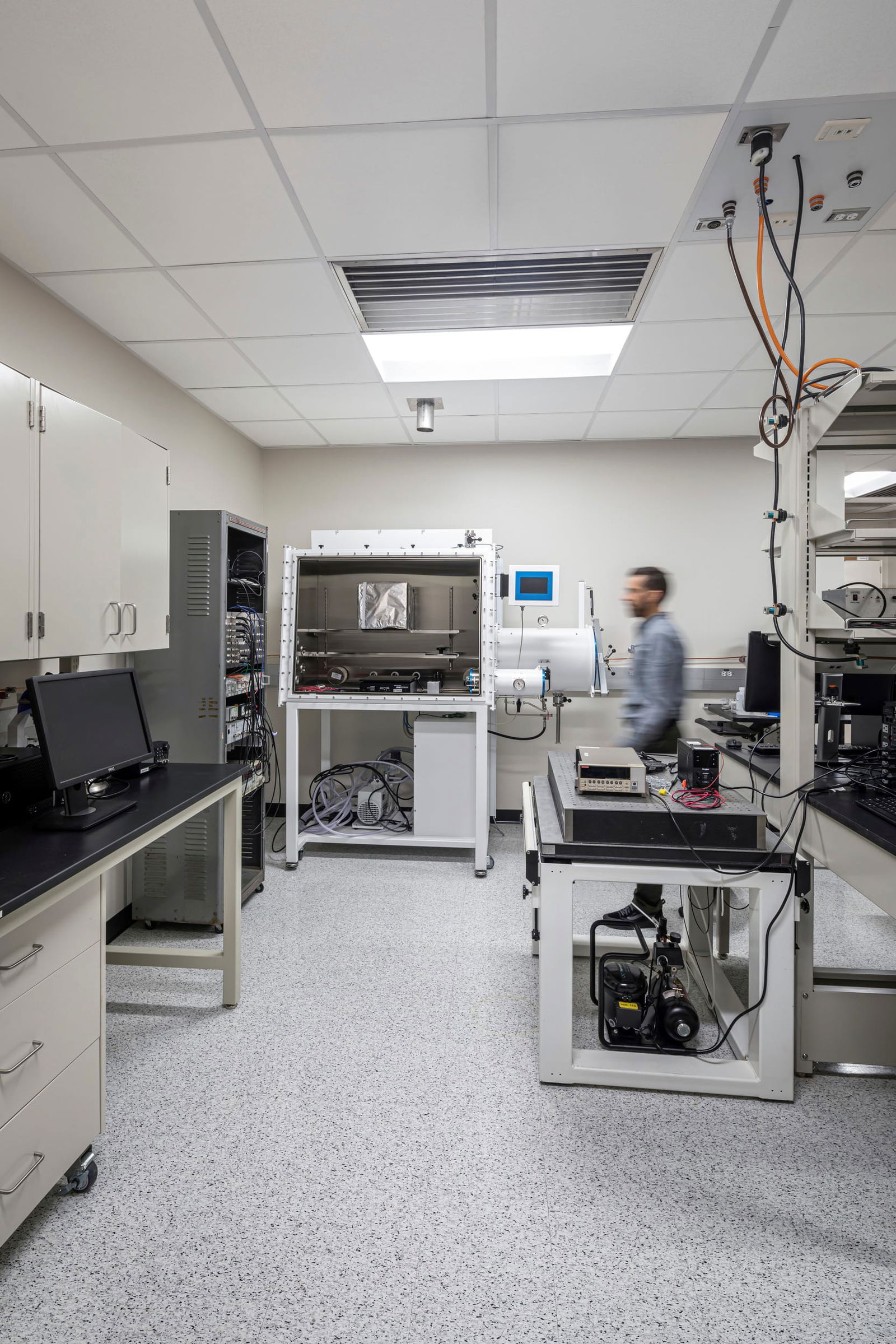 Advanced electrochemistry research laboratory at UT Austin designed by BSA featuring specialized equipment including a central environmental test chamber with digital control interface and vacuum capability. The lab includes bench-mounted measurement instruments, a computer workstation for data acquisition, and extensive power and gas connections dropping from the ceiling grid. A researcher in a light blue lab coat moves through the space equipped with white laboratory cabinetry, black countertops, speckled epoxy flooring, and ceiling-mounted utility services. The purpose-built facility houses vacuum pumps beneath the workbenches, precision electronic testing equipment, and environmental controls necessary for sensitive electrochemical experiments and material characterization.
