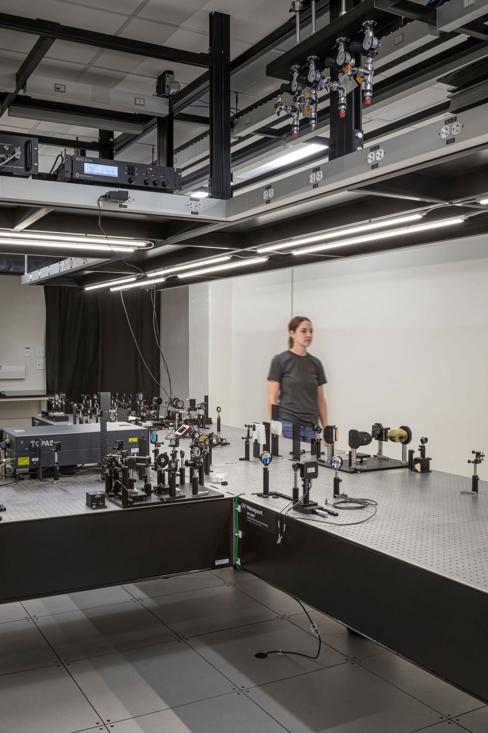 Advanced physics laboratory at UT Austin designed by BSA featuring optical breadboards populated with precision measurement equipment including mirrors, lenses, beam splitters, and laser components arranged on vibration-isolated tables. The space includes an overhead utility infrastructure with multiple gas valves, electrical connections, and control systems mounted on black metal framework with integrated LED lighting. A researcher wearing a gray t-shirt moves through the specialized research environment that features perforated optical tables, sophisticated testing apparatus, and blackout curtains for light-sensitive experiments. The lab's raised floor system and exposed ceiling design accommodate the complex requirements for physics research requiring exceptional stability and environmental control.
