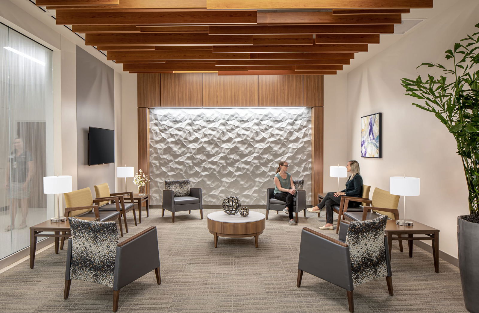 A modern healthcare waiting area with a contemporary design and warm feel. The space features a mix of seating options, including grey and black armchairs and wooden-framed lounge chairs. The walls are adorned with textured white paneling and warm wood accents, with a mounted flat-screen TV on one side. Hanging wooden beams decorate the entire ceiling, contributing to the warm and modern feel. Two women are seated in opposite chairs and are engaged in a conversation.
